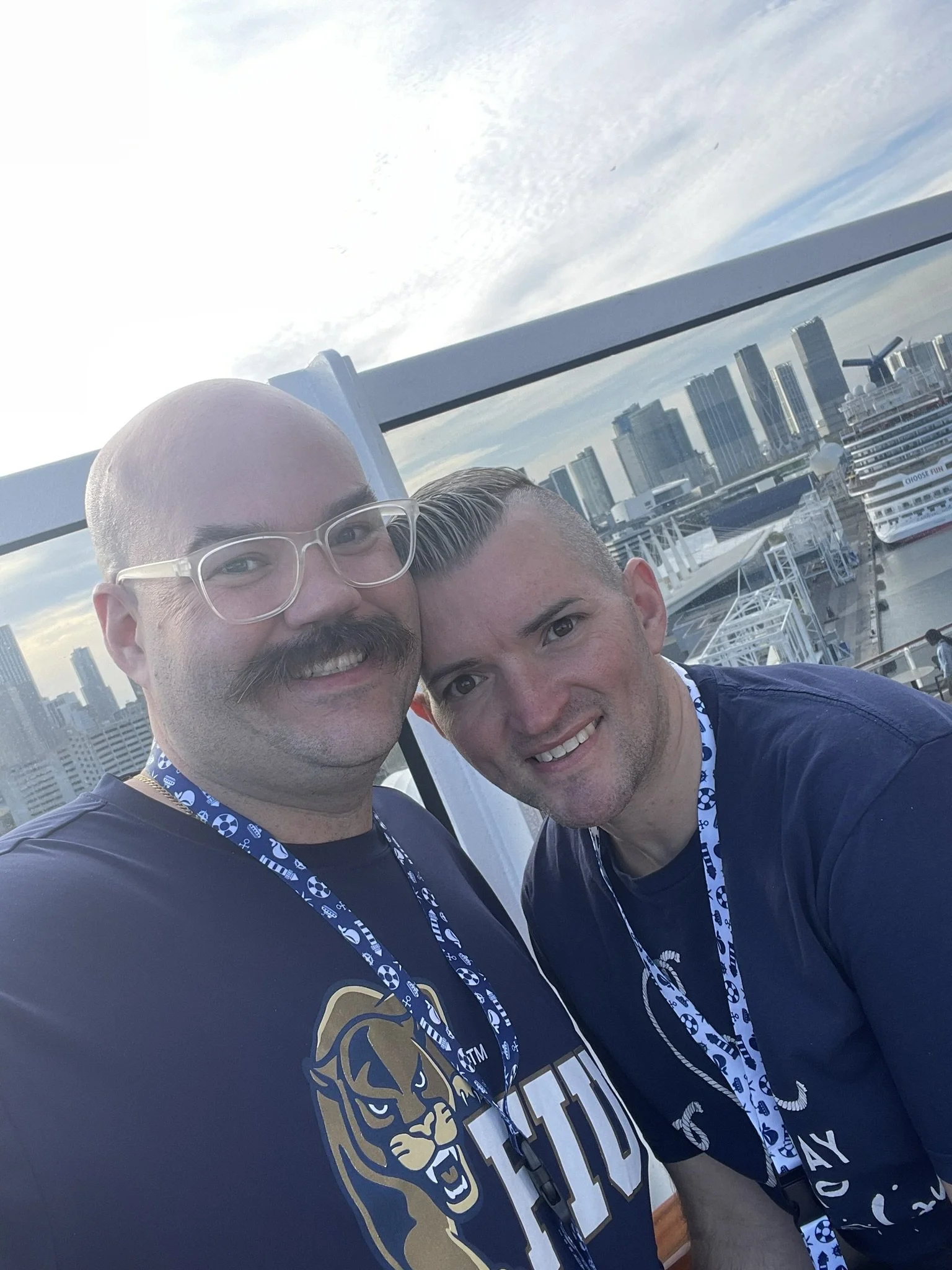 Two men smiling and posing for a selfie on a the top deck of a cruise ship with a city skyline and cruise ships in the background.
