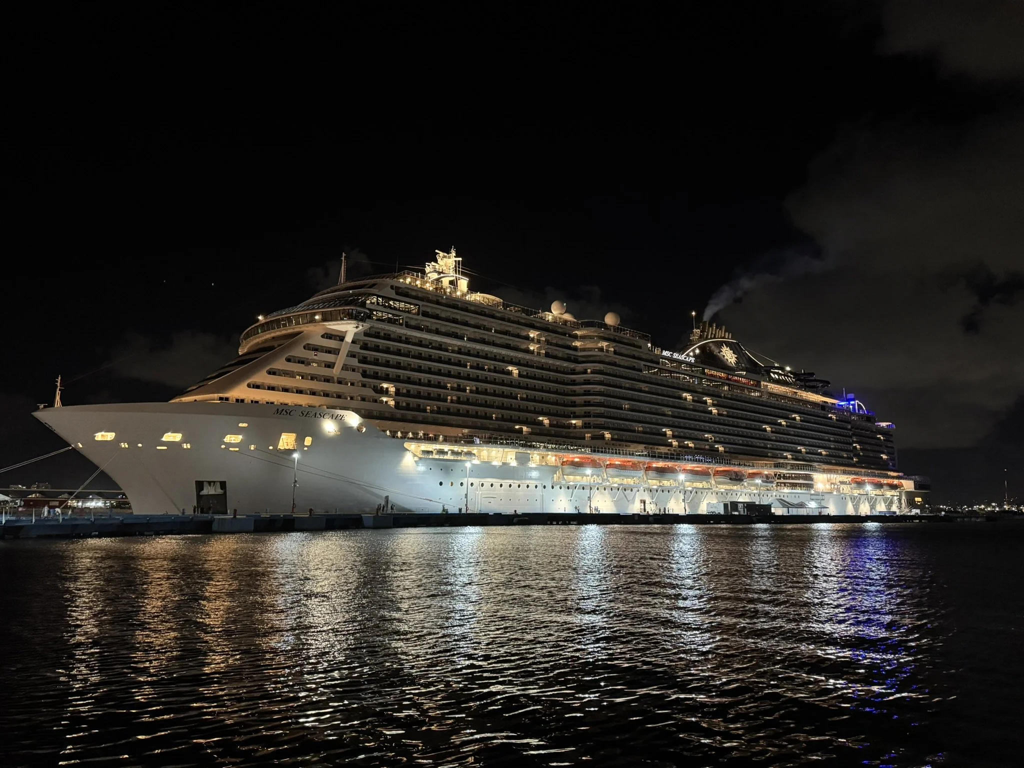 Night view of a large cruise ship named MSC Seascape docked at a port, illuminated with lights reflecting on the water.