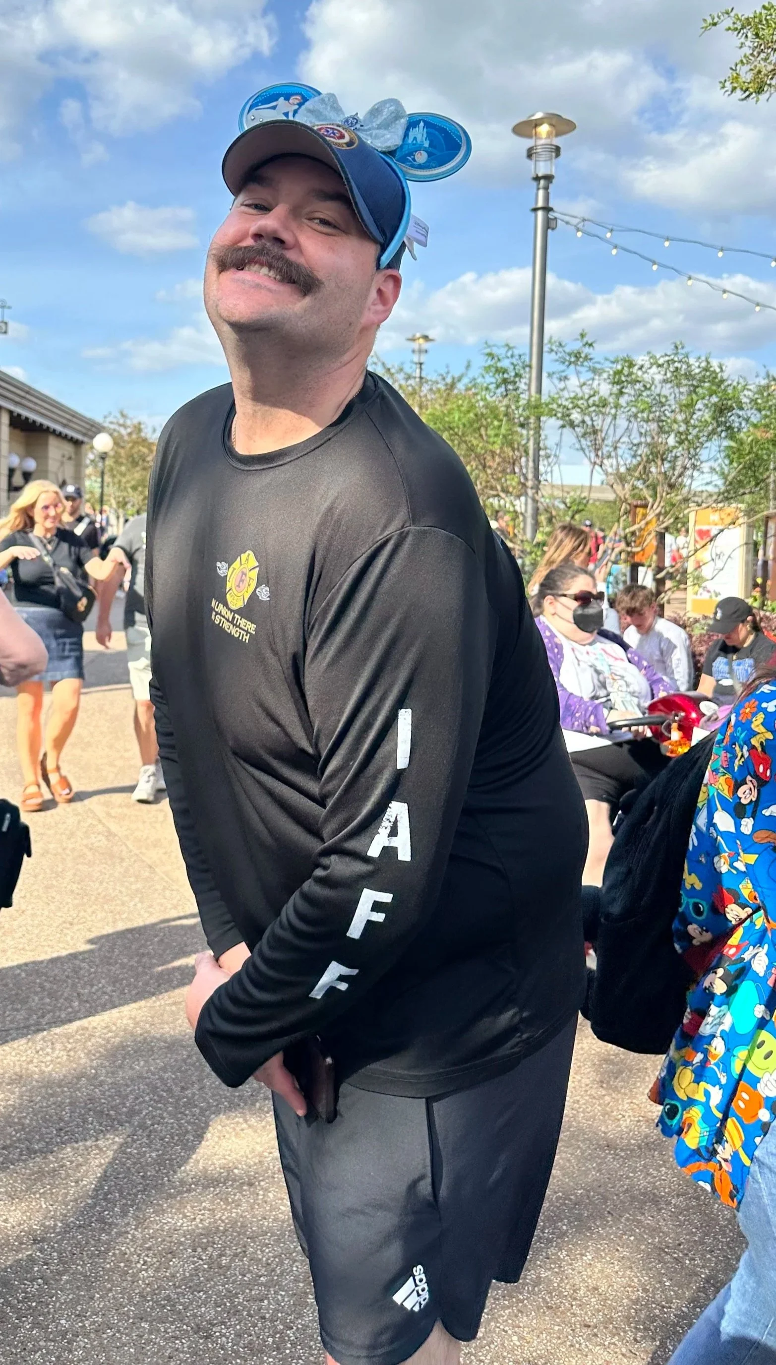 A man with a mustache smiling at the camera, wearing Mickey Mouse ears with a blue cap, a black long-sleeve shirt with