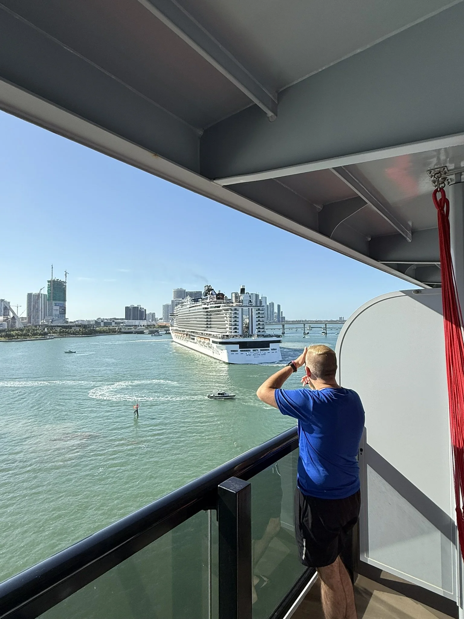 A man in a blue shirt and black shorts on a balcony watching a large cruise ship pass by on a river with city skyscrapers in the background.