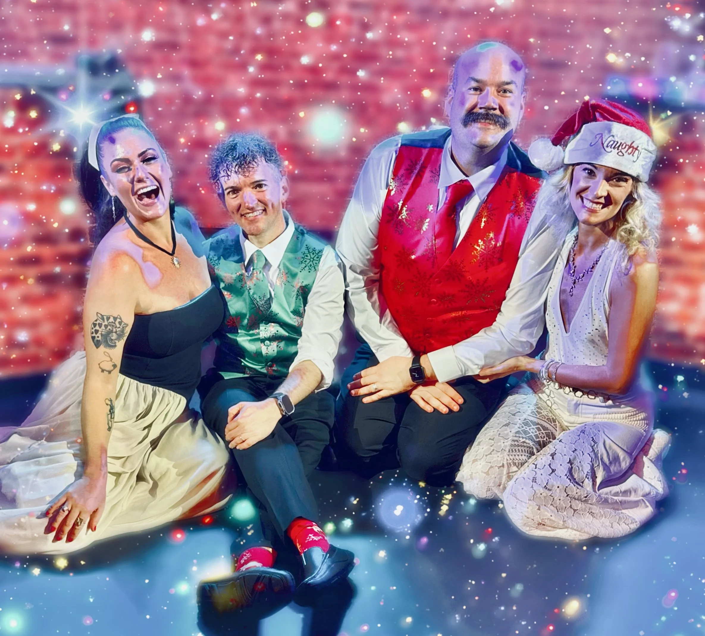 Group of four people in festive holiday attire celebrating Christmas, sitting on the floor in front of a red brick wall, with sparkles and lights added for a cheerful effect.