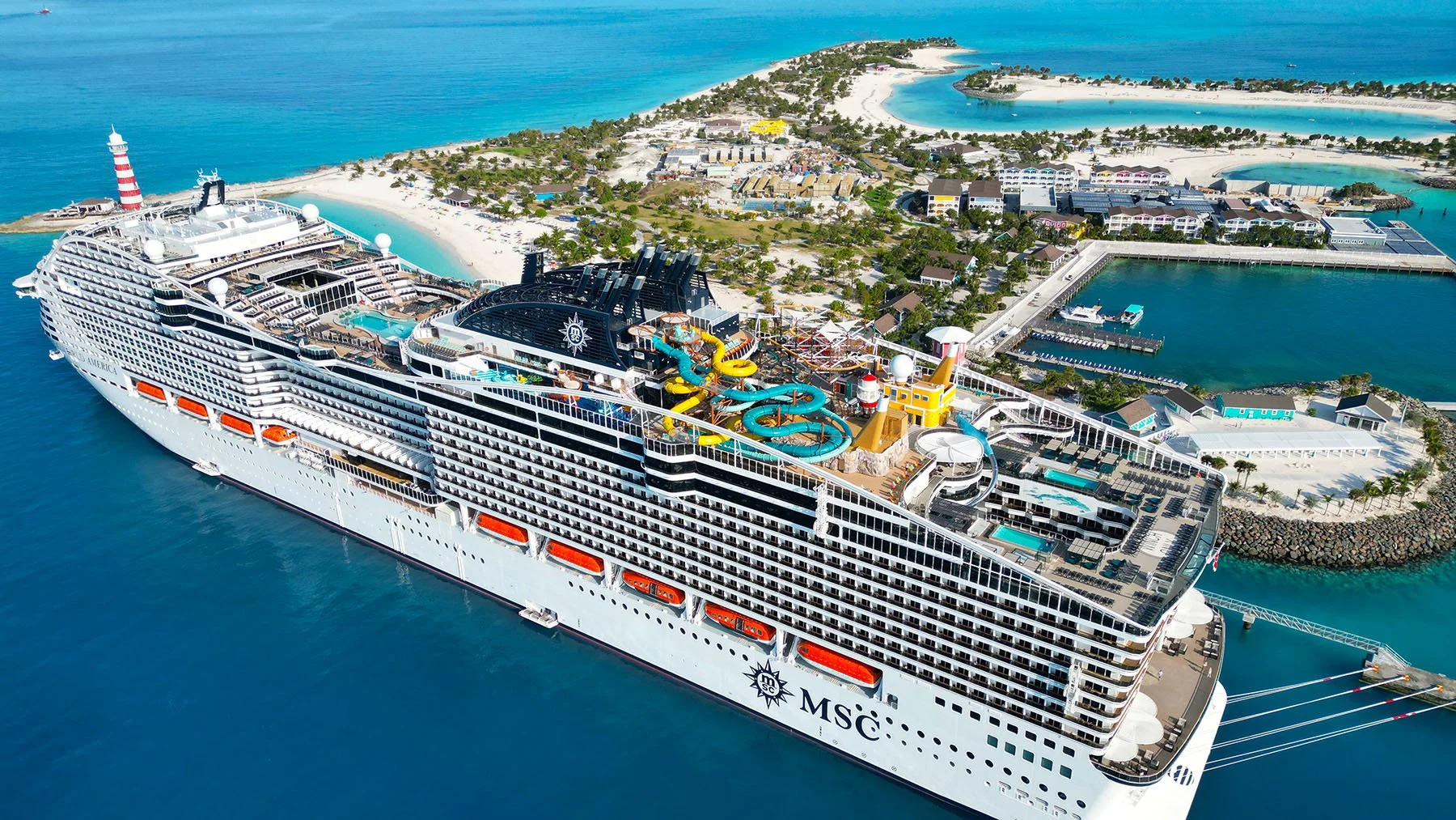 A large cruise ship docked near a tropical island with white sandy beaches, water parks, pools, and lush green vegetation.