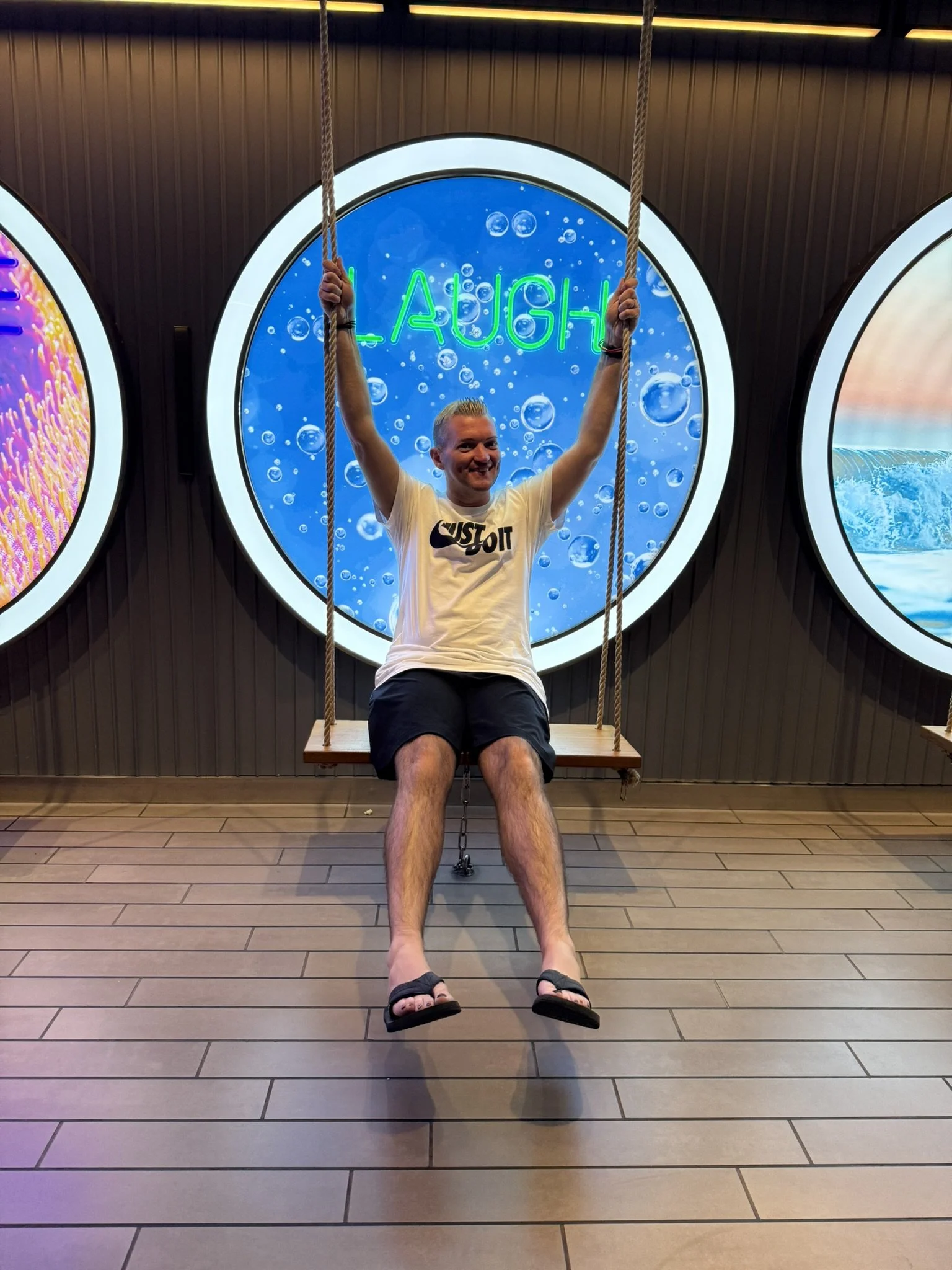 A man sitting on a swing with his arms raised, smiling, in front of a blue circular background with the word 'LAUGH' in neon green. The man is wearing a white T-shirt with a Nike logo and text, black shorts, and flip-flops.