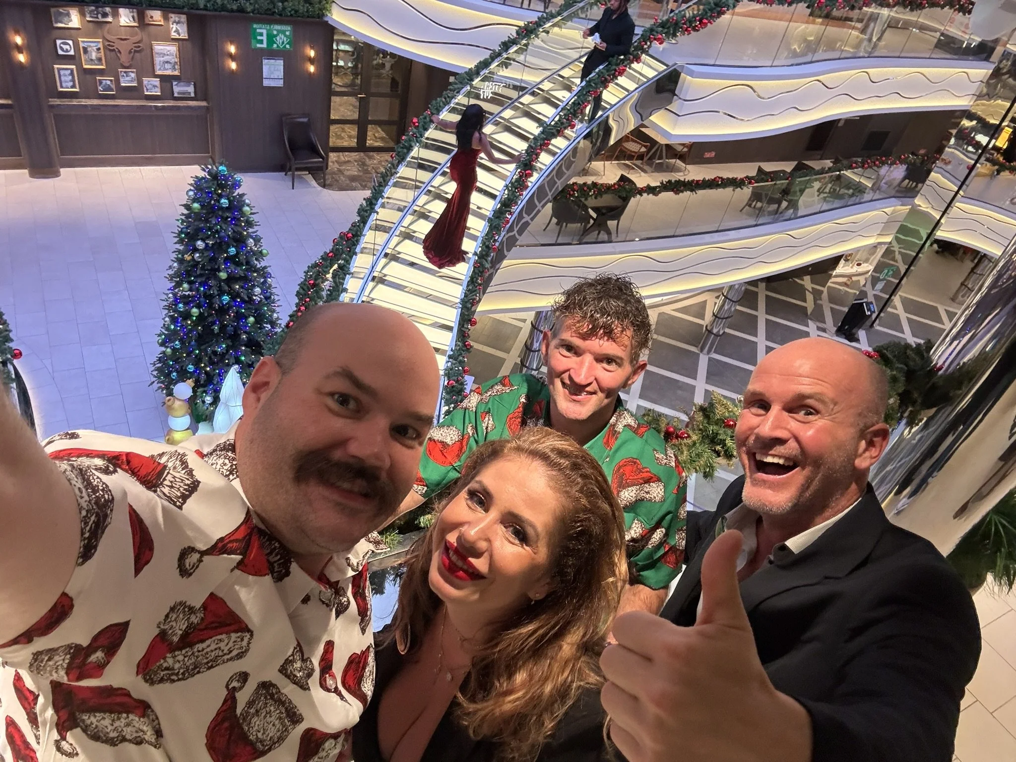 A group of four people celebrating Christmas inside a decorated shopping mall or hotel, taking a selfie with a Christmas tree, garlands, and a Christmas-themed woman in a red dress on the escalator in the background.