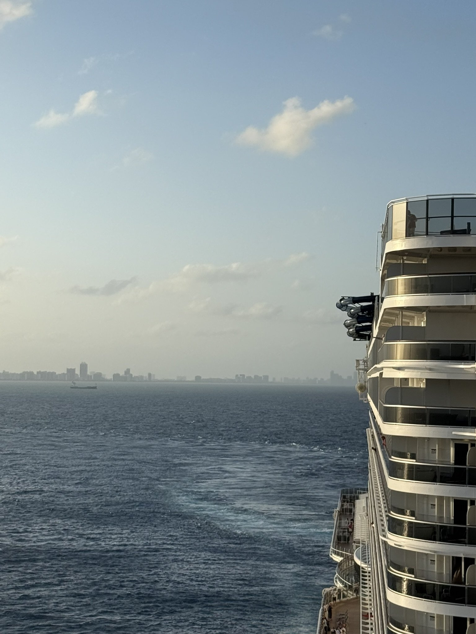 A view from a cruise ship showing the ocean with a city skyline in the distance, part of the ship's side with balconies, and a clear sky with some clouds.