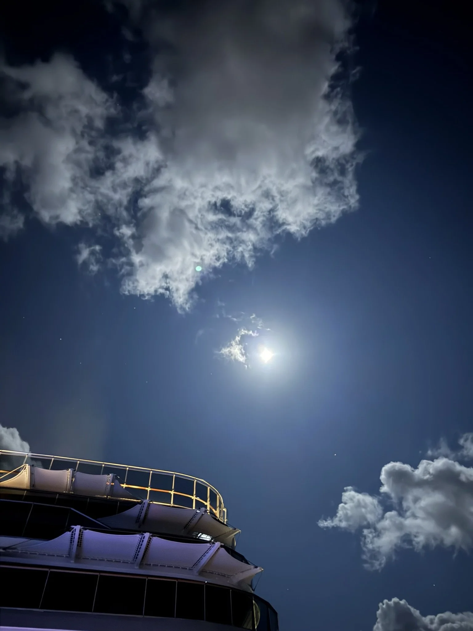 Night sky with clouds, bright moon, and visible stars above a building with a railing.