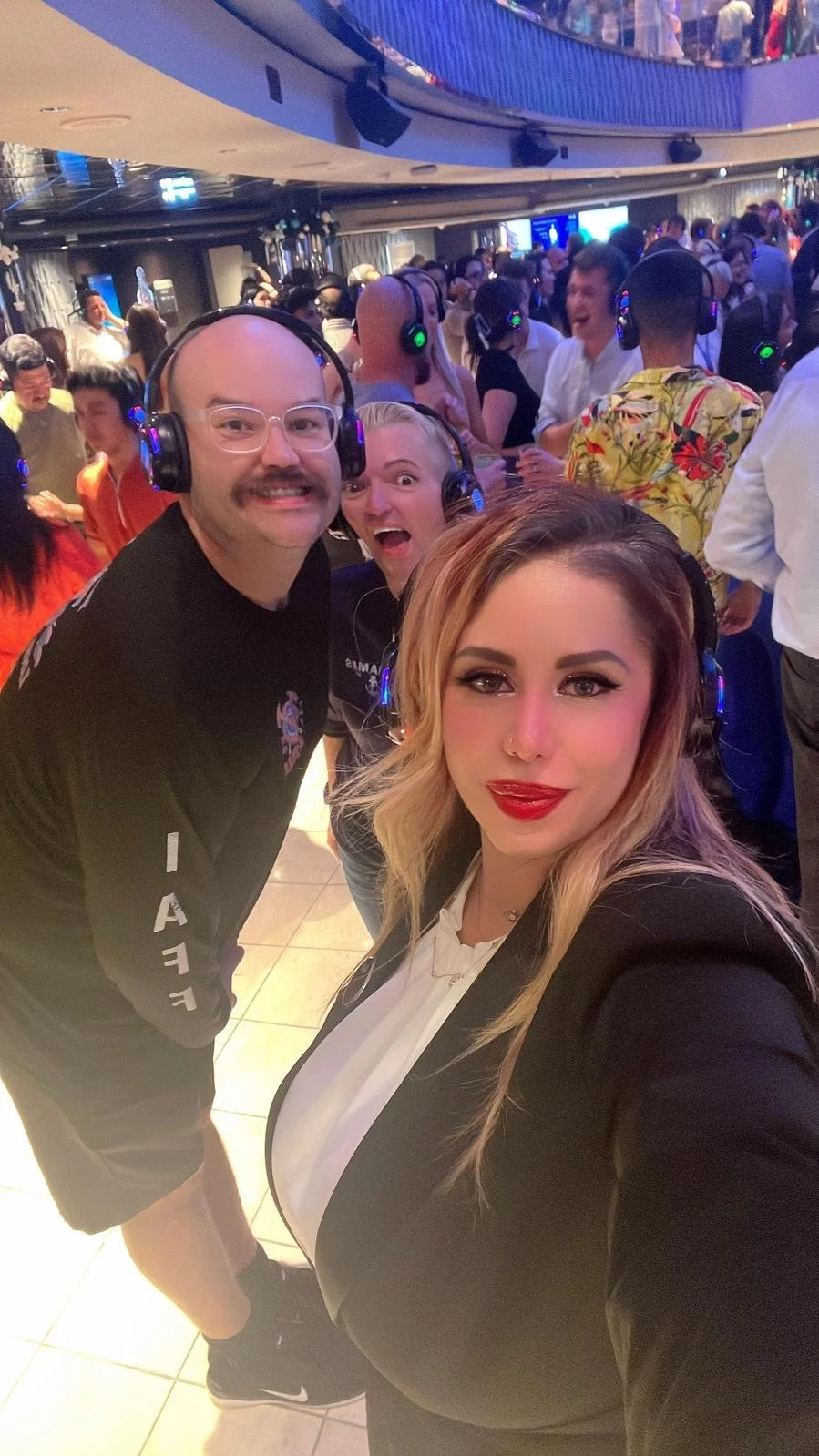 Three people wearing headphones taking a selfie at a crowded party or event with many other people in the background.
