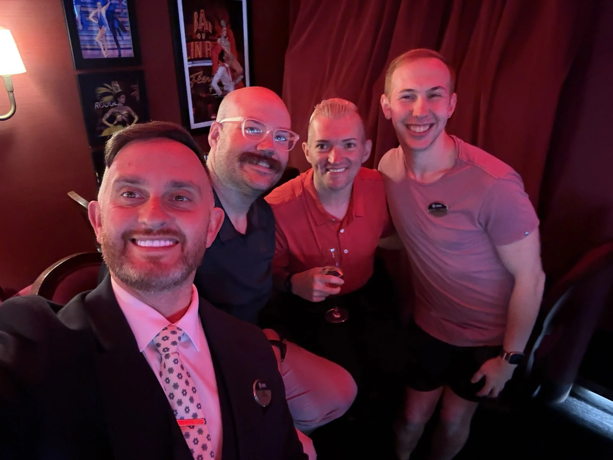Four men smiling and posing together in a dimly lit room with red lighting and posters on the wall.