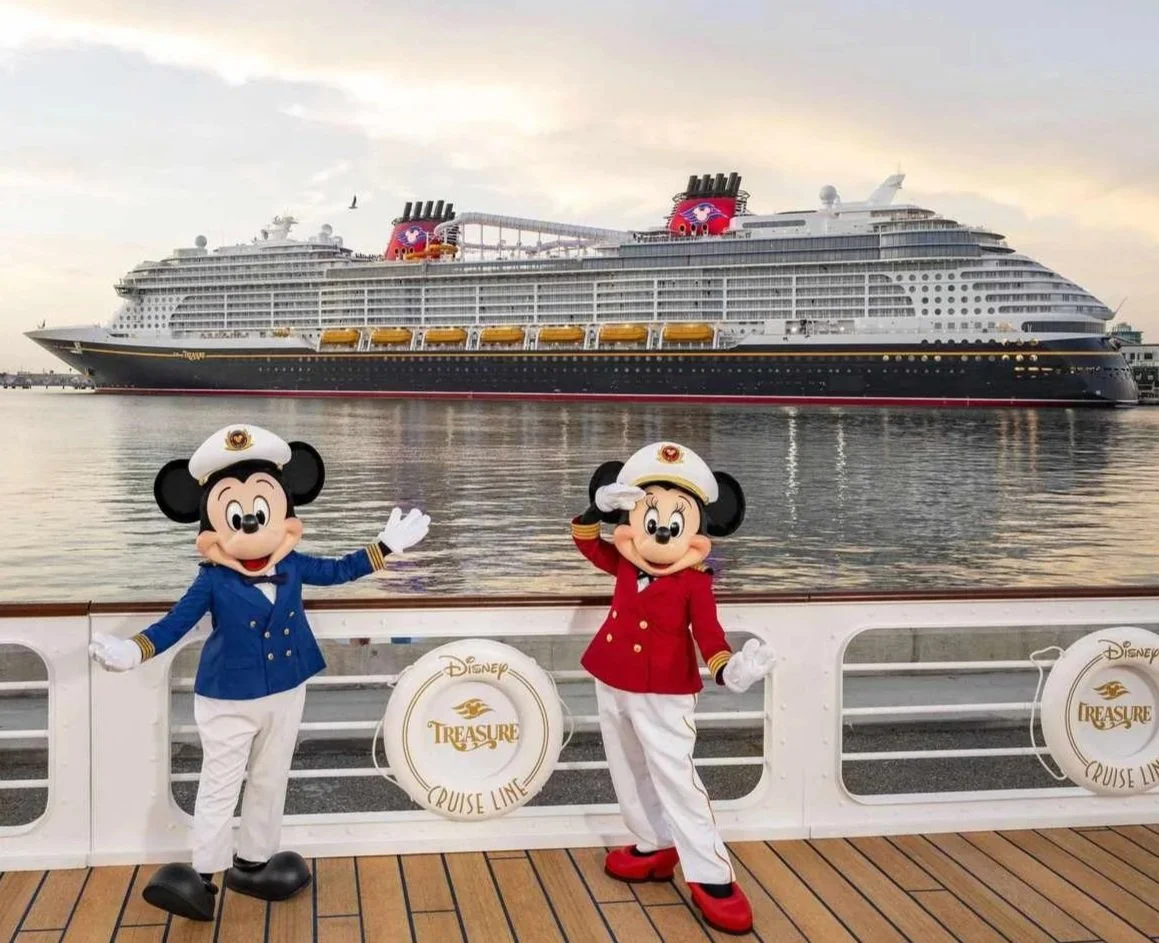 Mickey Mouse and Minnie Mouse dressed as captains on the deck of a cruise ship with the Disney Treasure in the background.