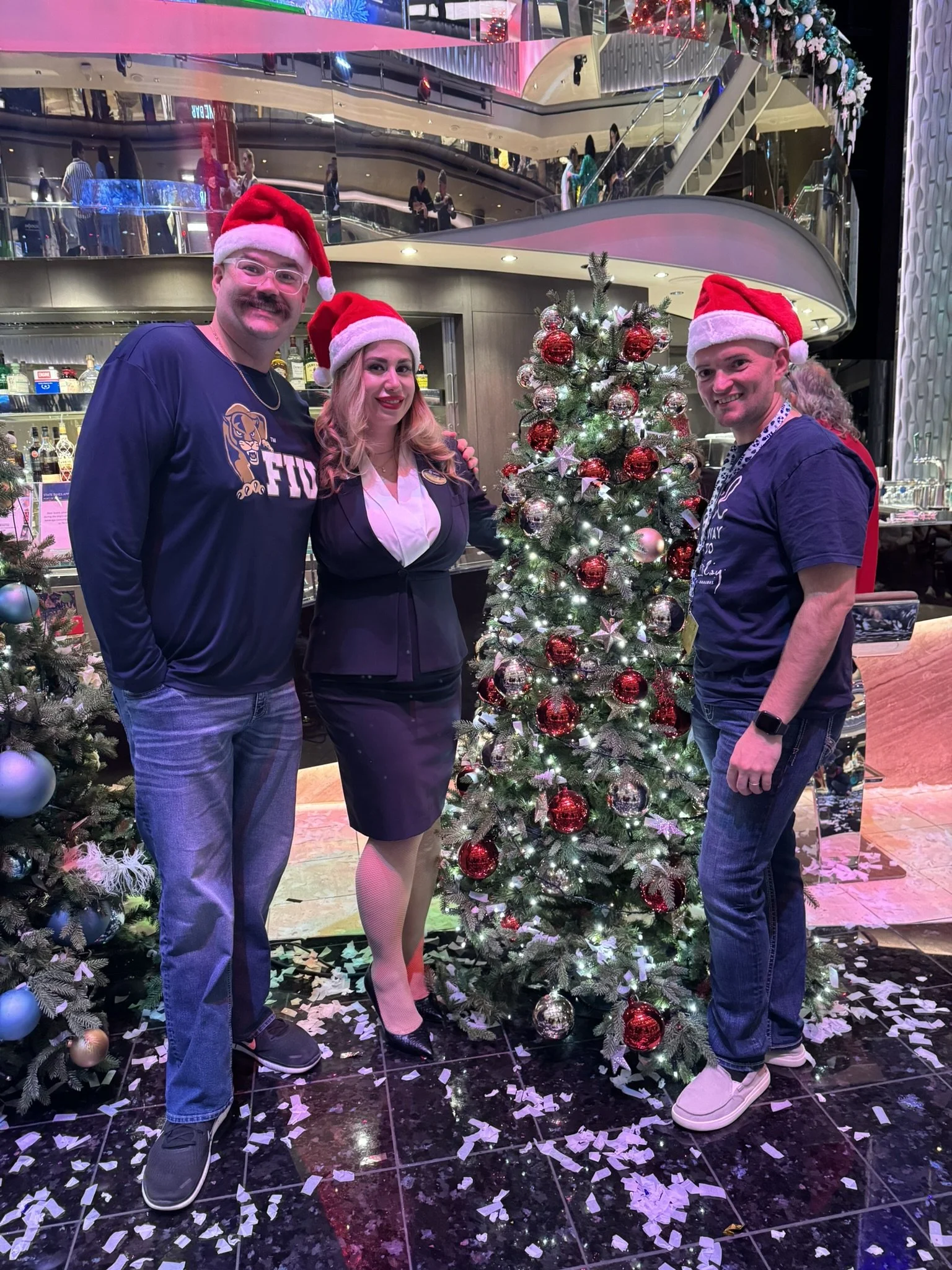 Three people wearing Santa hats posing next to a decorated Christmas tree in a festive indoor setting.