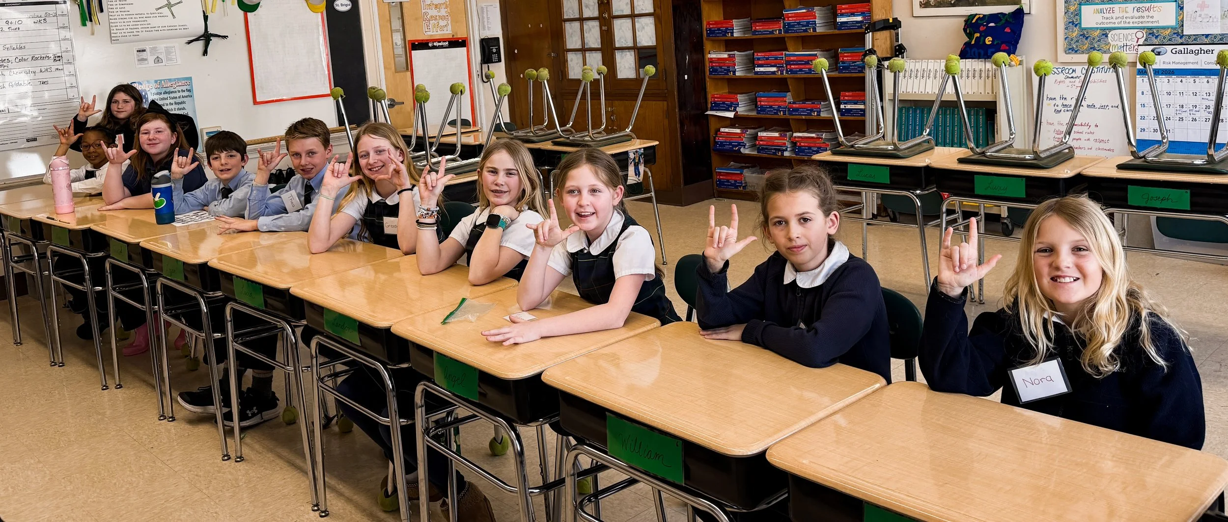 A classroom with ten children sitting at desks, making the sign of the horns with their hands, some smiling and some with serious expressions. The children are wearing school uniforms and name tags, and the classroom has bookshelves and desks with chairs stacked upside down.