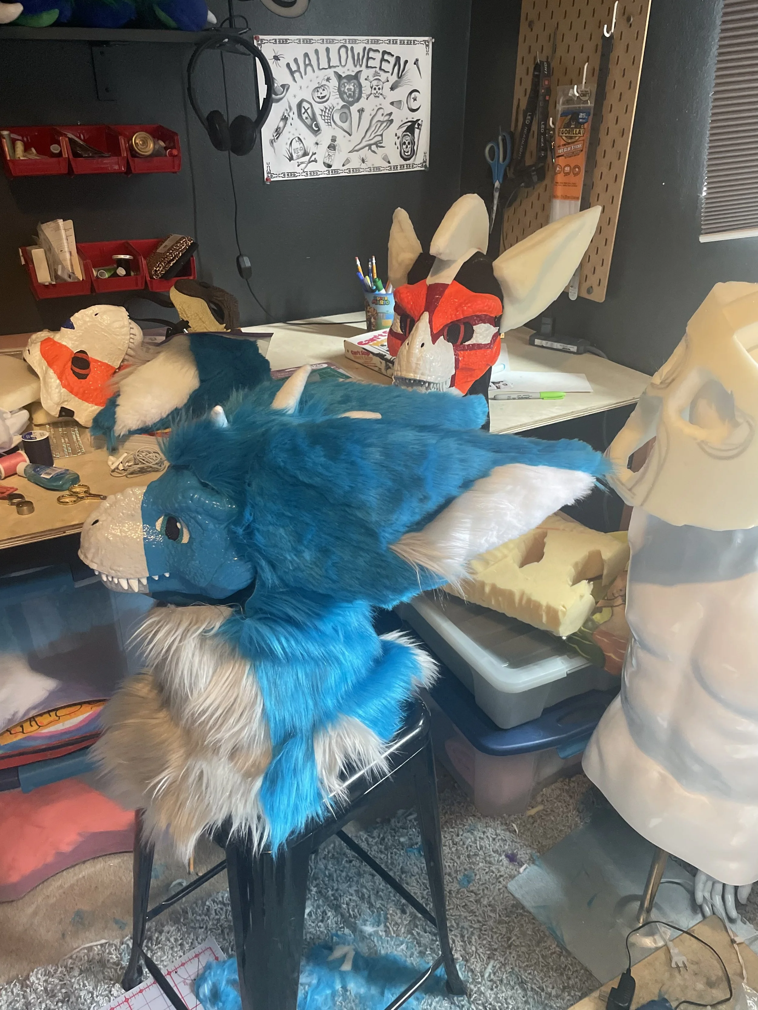 A workspace with various costume parts, including a blue furry dragon head with large horns, white teeth, and detailed eyes, alongside a red and black fox mask, and a white skull mask. The background includes a Halloween-themed poster, crafting supplies, scissors, markers, and foam materials, indicating it's a costume-making area.