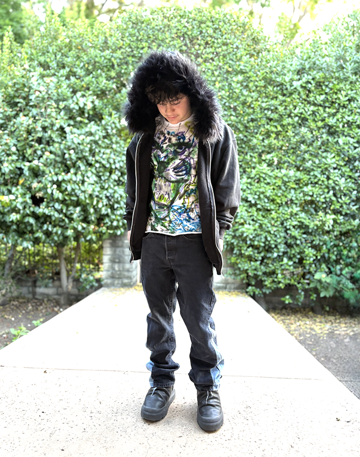 Young person standing on outdoor concrete walkway, wearing a black fur-lined hooded jacket, multicolored graphic T-shirt, black pants, and black boots, with greenery in the background.