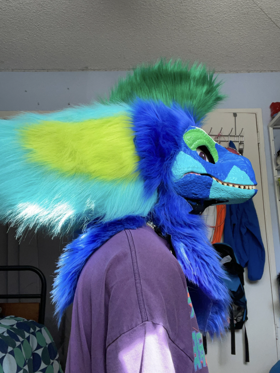 A person wearing a colorful dragon or creature costume with a large blue face, green, blue, and yellow fur details, and a purple shirt in a room with a bed, lockers, and clothing in the background.