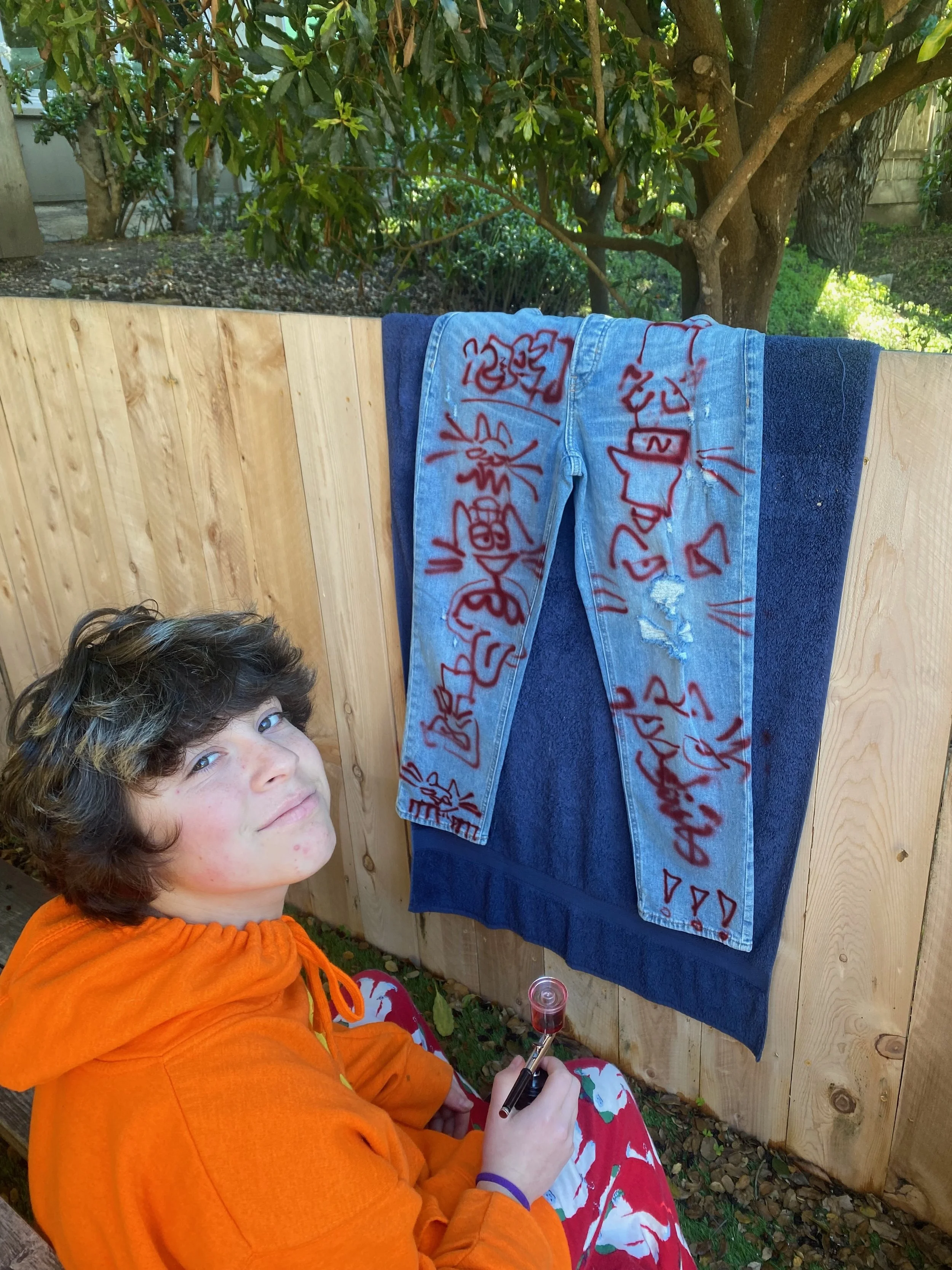 A young boy with curly hair, wearing an orange hoodie and red- and white patterned sweatpants, sitting outdoors near a wooden fence. He is holding a marker and appears to be drawing or writing on a pair of jeans hanging on the fence. The jeans are de