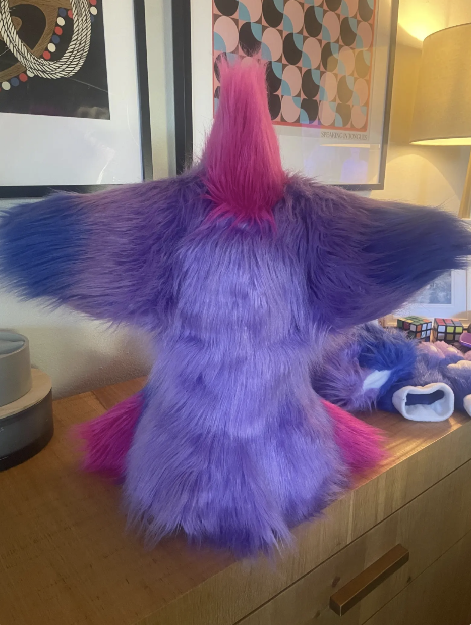 Colorful furry sculpture resembling a mythical creature with a pink horn, purple body, and pink tail, sitting on a wooden surface in a room with framed artwork and a lamp.