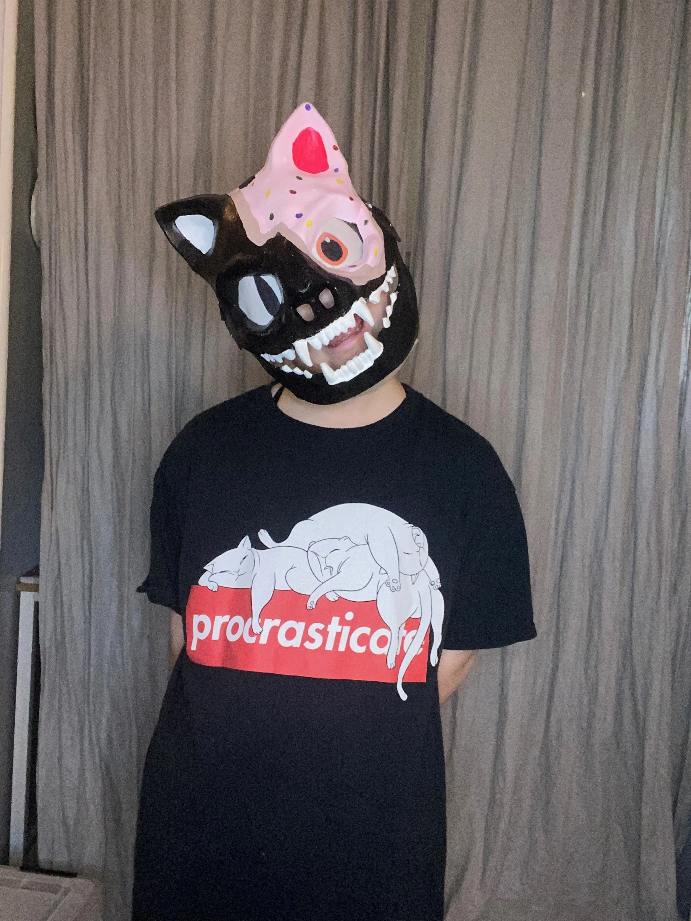 Person wearing a black mask designed as a cat with a painted face mask resembling a pig, standing in front of a grey curtain, wearing a black t-shirt with a cat illustration and the word "procrastinates" on it.