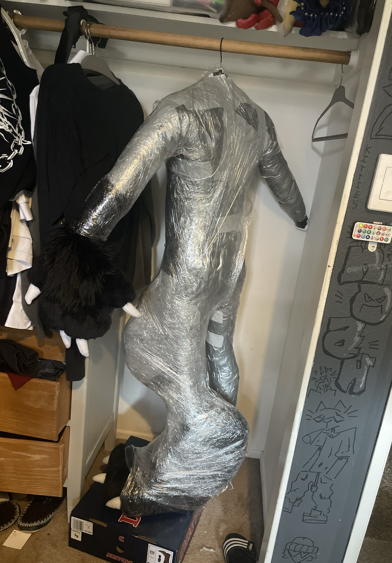 A mannequin wrapped in clear plastic wrap stands inside a closet, surrounded by hanging clothes and drawers. To the right, there is a partial chalkboard wall with doodles and writing, and a remote control is on a nearby surface.