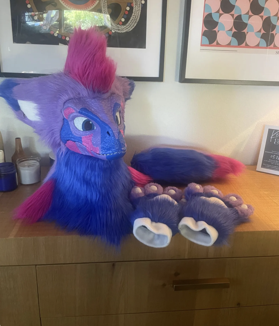 Colorful anthropomorphic creature costume head with purple fur, pink mohawk, and blue face with pink and purple markings, along with matching paws and a multicolored tail, placed on a wooden surface in a room with framed artwork on the wall.
