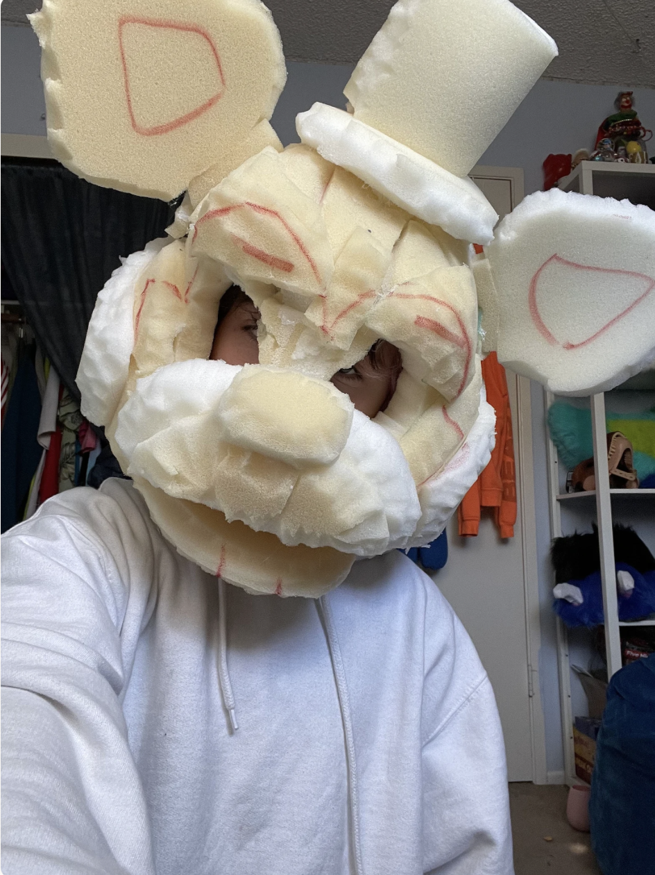 Person wearing a large, homemade mask resembling Santa Claus made of foam and padding in a cluttered room.