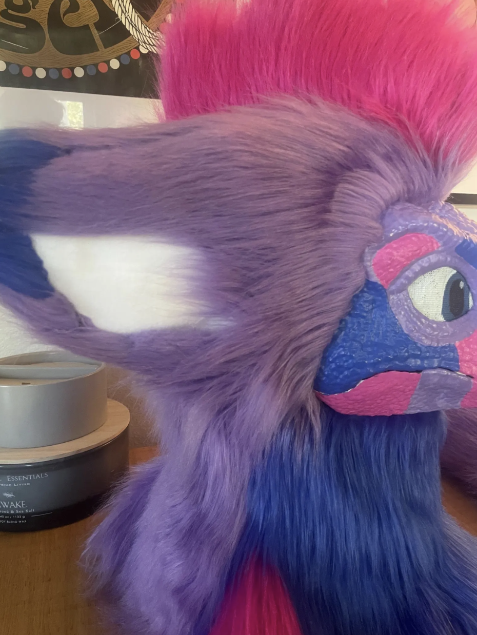 Close-up of a colorful plush toy with purple, pink, blue, and white fur, and a painted face with large eyes, on a wooden surface next to a candle and a frame.
