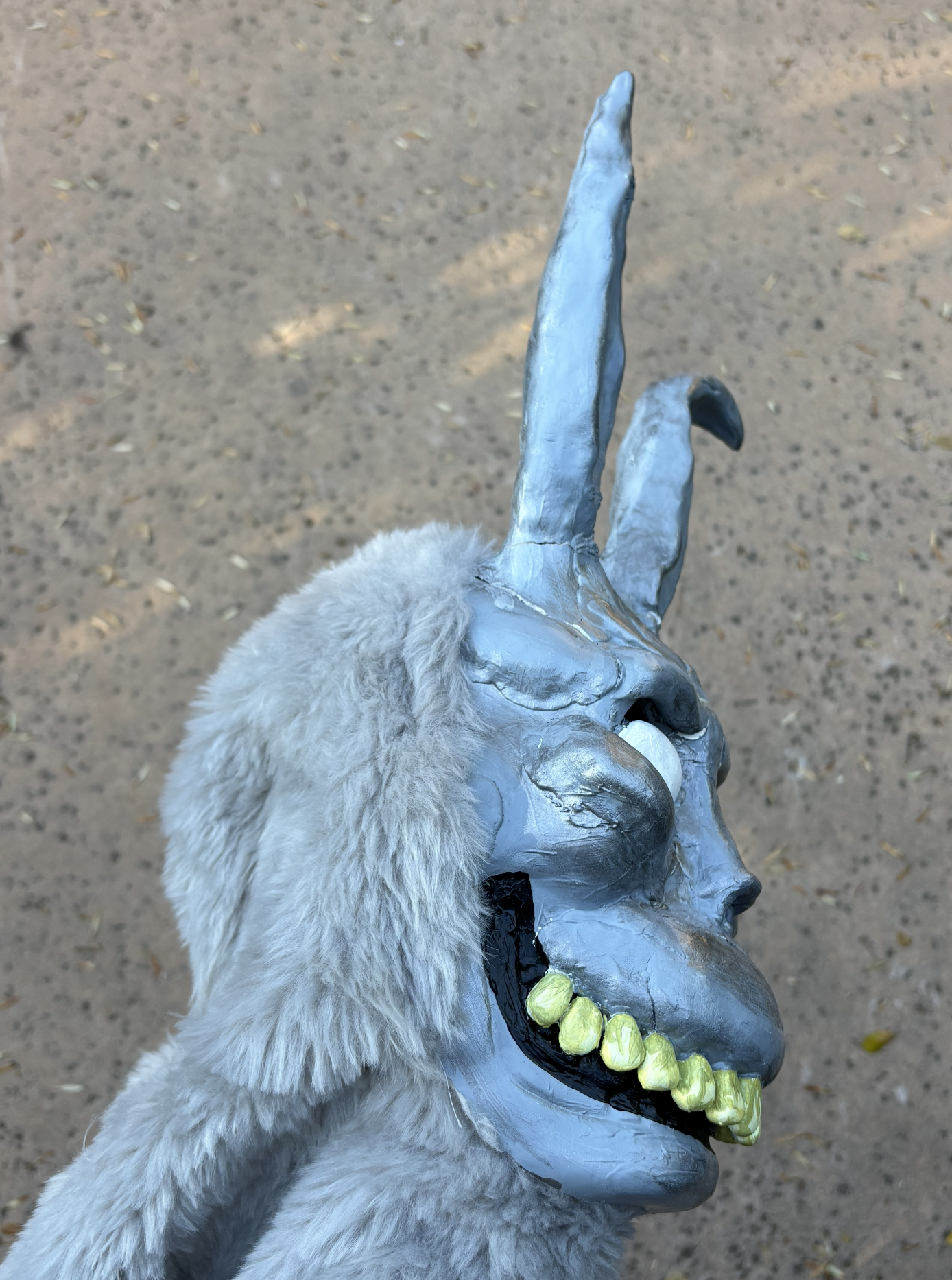A hand covered in a shiny silver glove making a peace sign. The glove features three long, pointed fingers and one slightly shorter finger, resembling a stylized animal or creature. The background shows a dirt ground with scattered leaves.