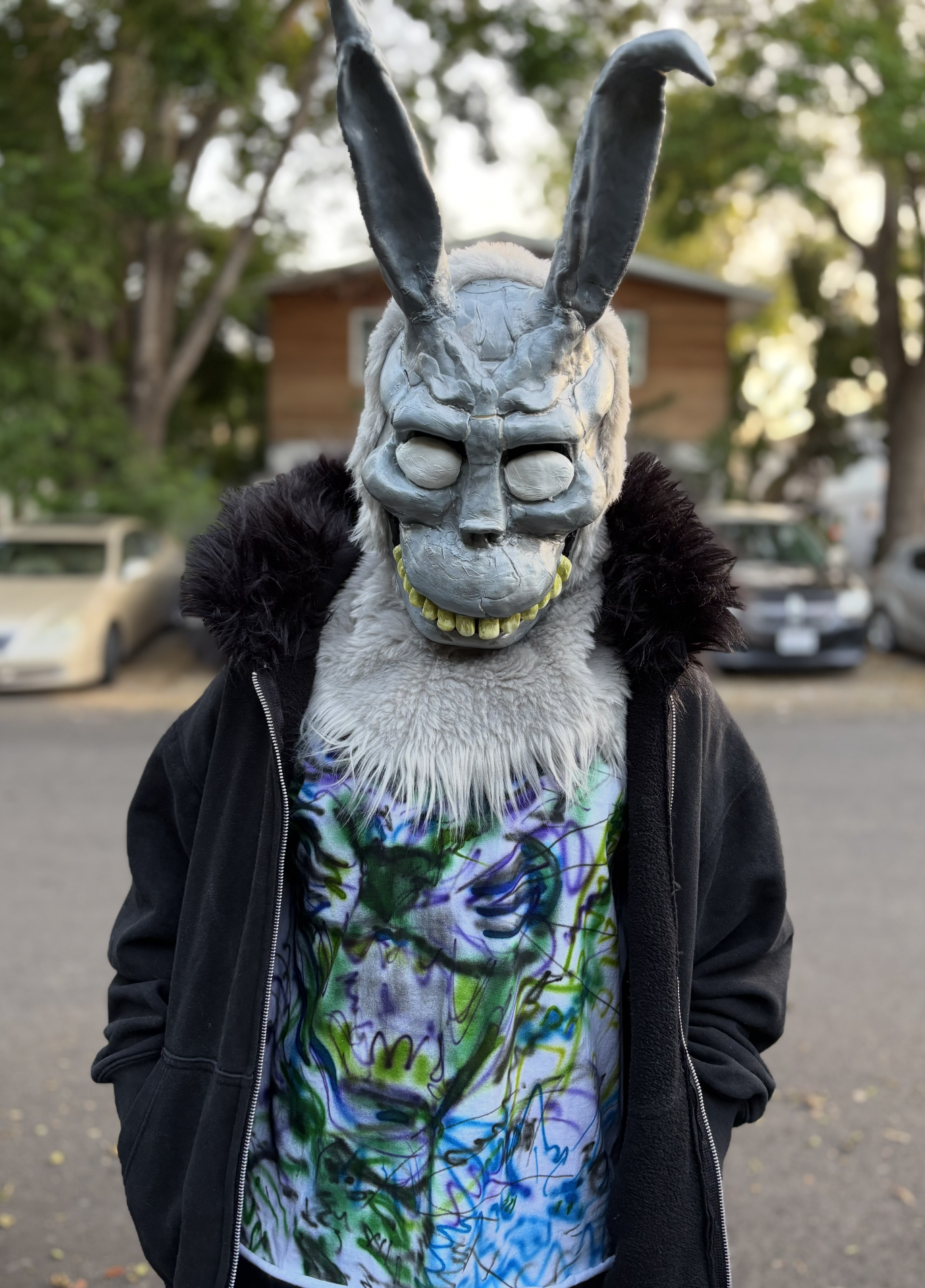 Person wearing a skull mask with rabbit-like ears, a black jacket, and colorful tie-dye shirt, standing outdoors with trees and houses in the background.