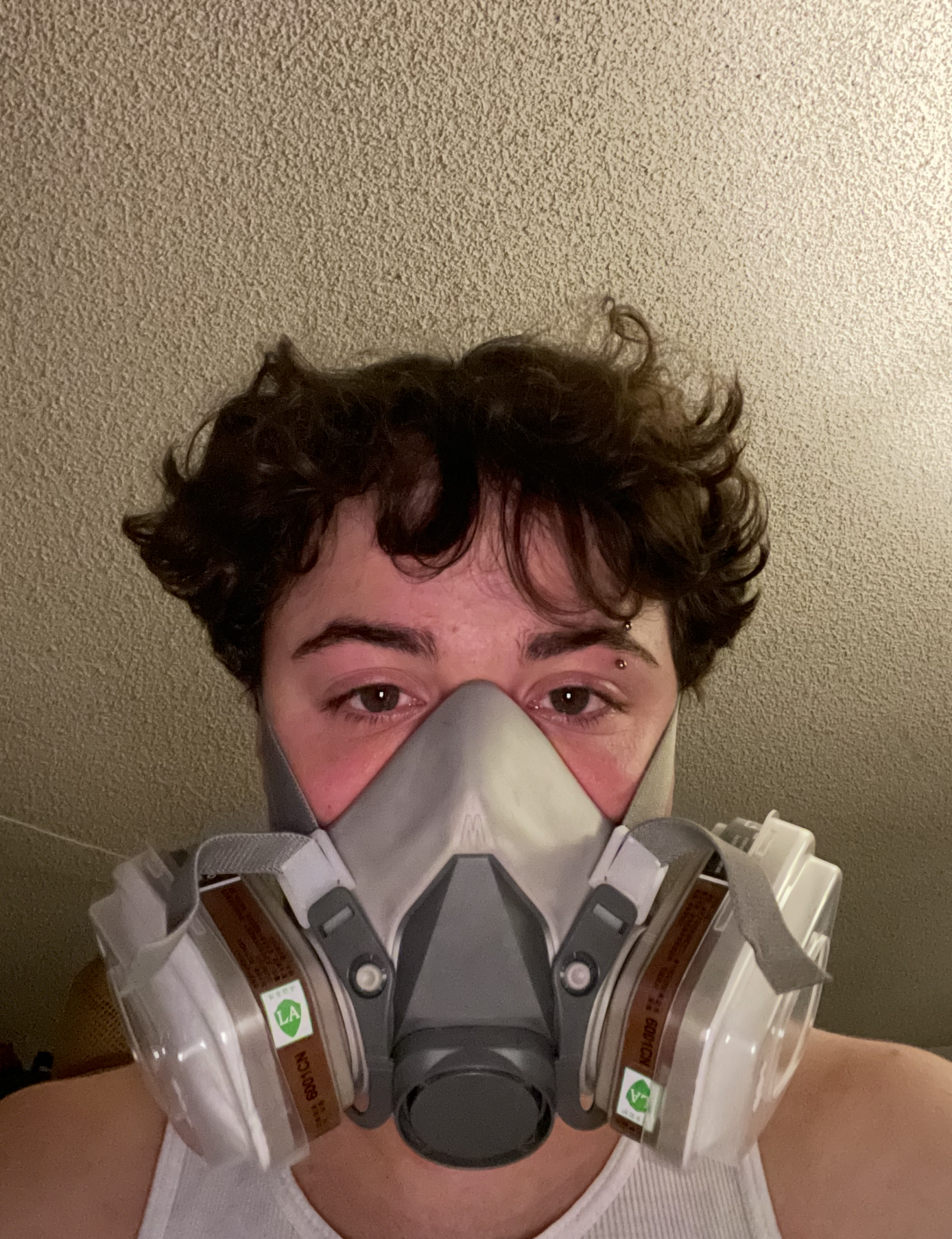 A person with curly hair is wearing a respirator mask and a white tank top, standing against a beige textured wall.