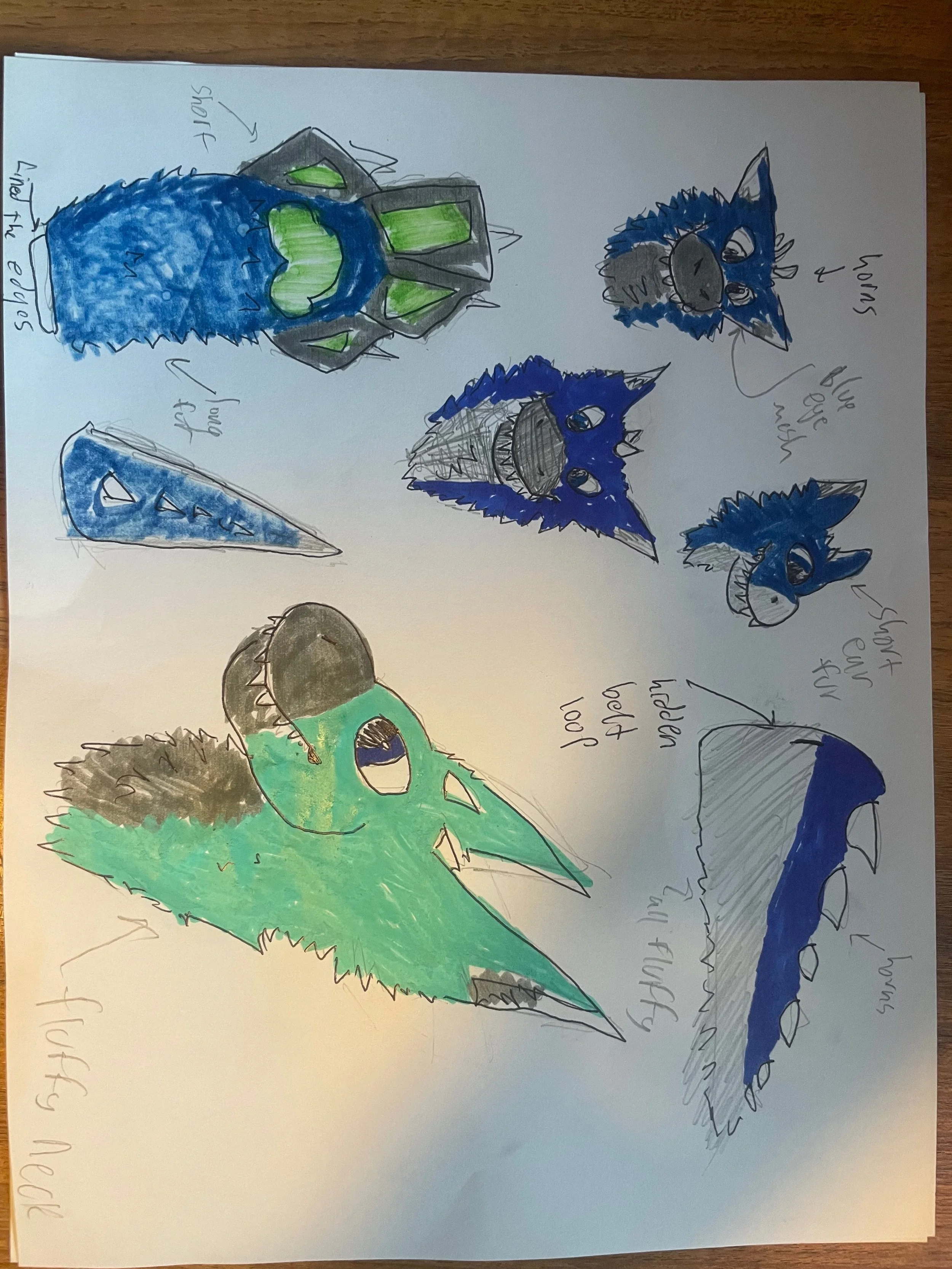 A sheet of paper with hand-drawn illustrations of various cartoon dinosaur heads, colored in shades of blue, green, and gray, with handwritten notes and labels pointing out features such as spikes, scales, and teeth.