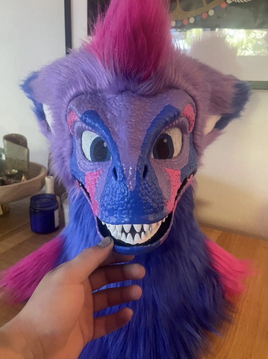 A colorful dragon mask with purple, pink, and blue hues, featuring large eyes, sharp teeth, and textured skin, being held in front of a furry costume body with matching colors.