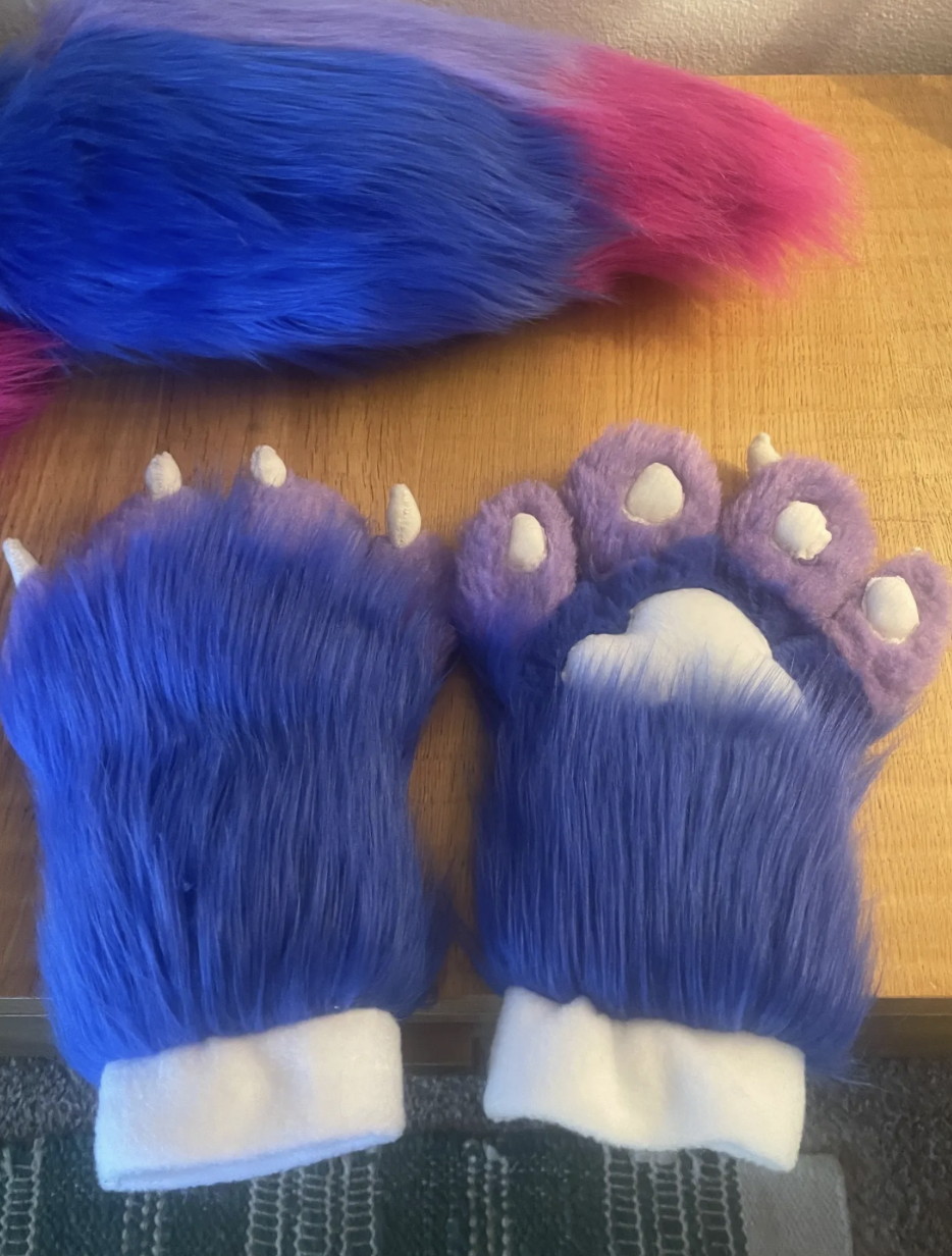 Colorful unicorn-themed pet costume set with a blue tail, a purple paw mitt with a white paw print, and a matching blue leg cover with white cuffs.