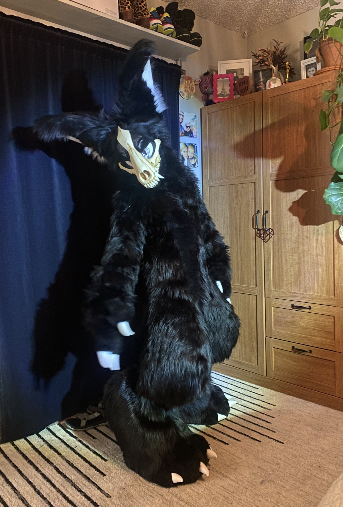 A person in a black furry costume with white claws and a skull mask is holding two large black stuffed animals resembling wolves, one is sitting on the floor and the other is being held.