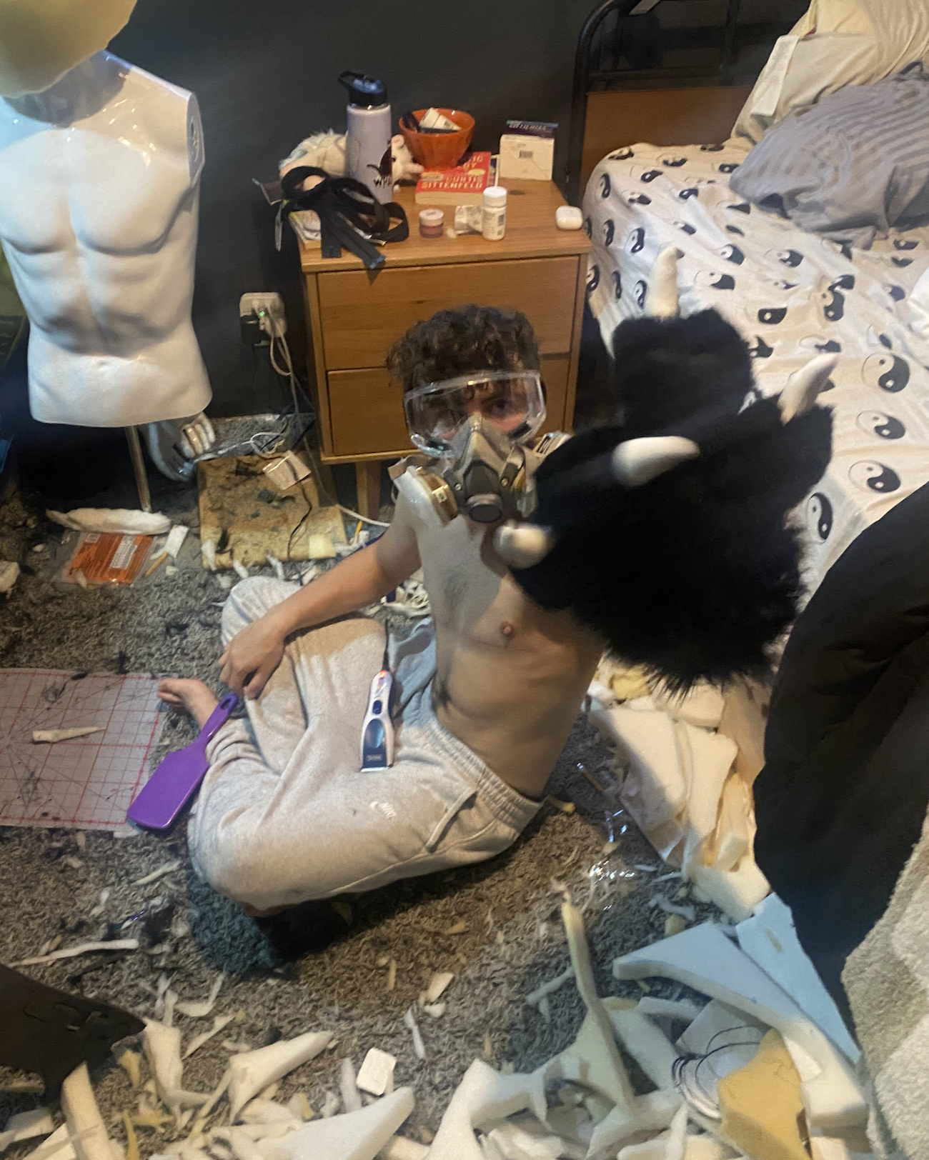 A young man wearing a gas mask and goggles, lying on the floor with a toy lightsaber, surrounded by debris including shredded paper, with a black and white puppy sitting on his chest, in a cluttered bedroom.
