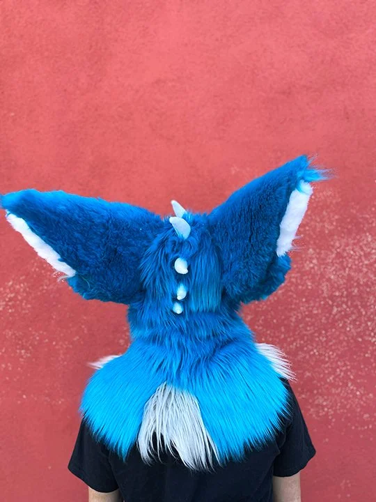 BACK OF HEAD: 
Dragon character rear view. This view is showing the big ears, and different lengths of faux fur while standing against a red textured wall.
(Materials used: Faux fur, acrylic paint, fleece, hand stitching and use of sewing machine)