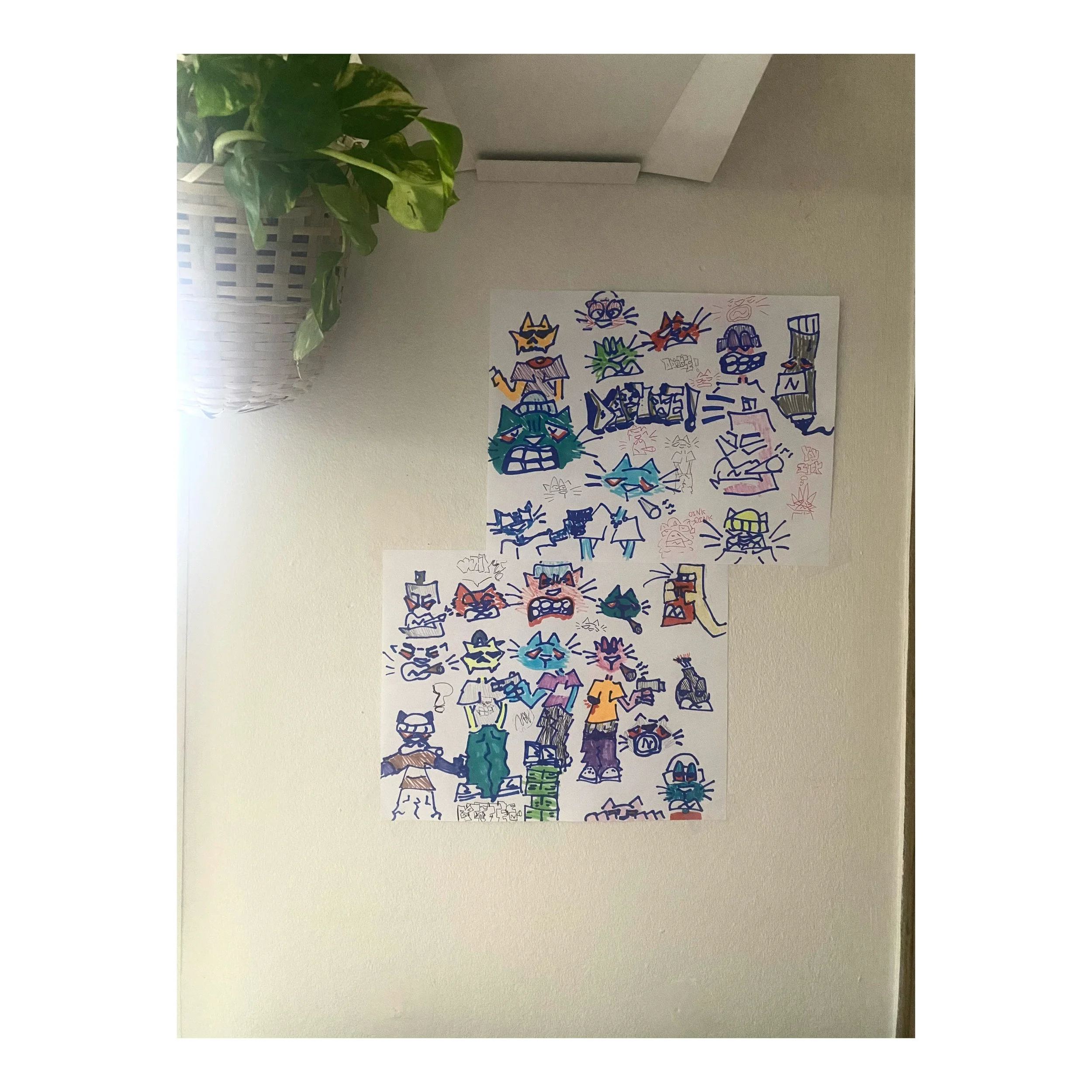 Colorful drawings of cats with expressive faces and various accessories, arranged on two sheets of paper taped to a beige wall, with a potted plant with green leaves in the top left corner.