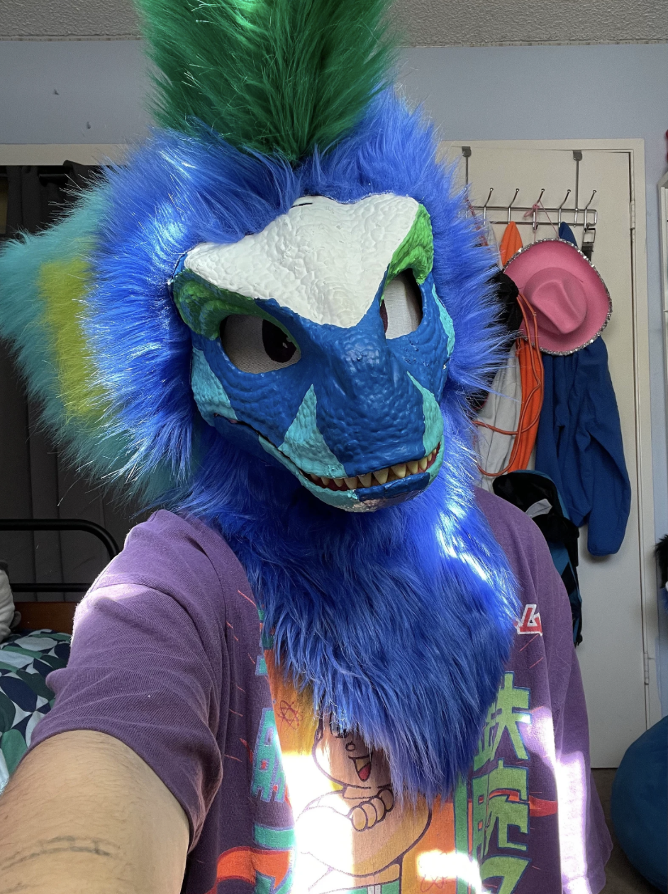 Person wearing a bright blue and green lion mask with colorful fur, taking a selfie in a room with various hats and clothing hanging on a wall.