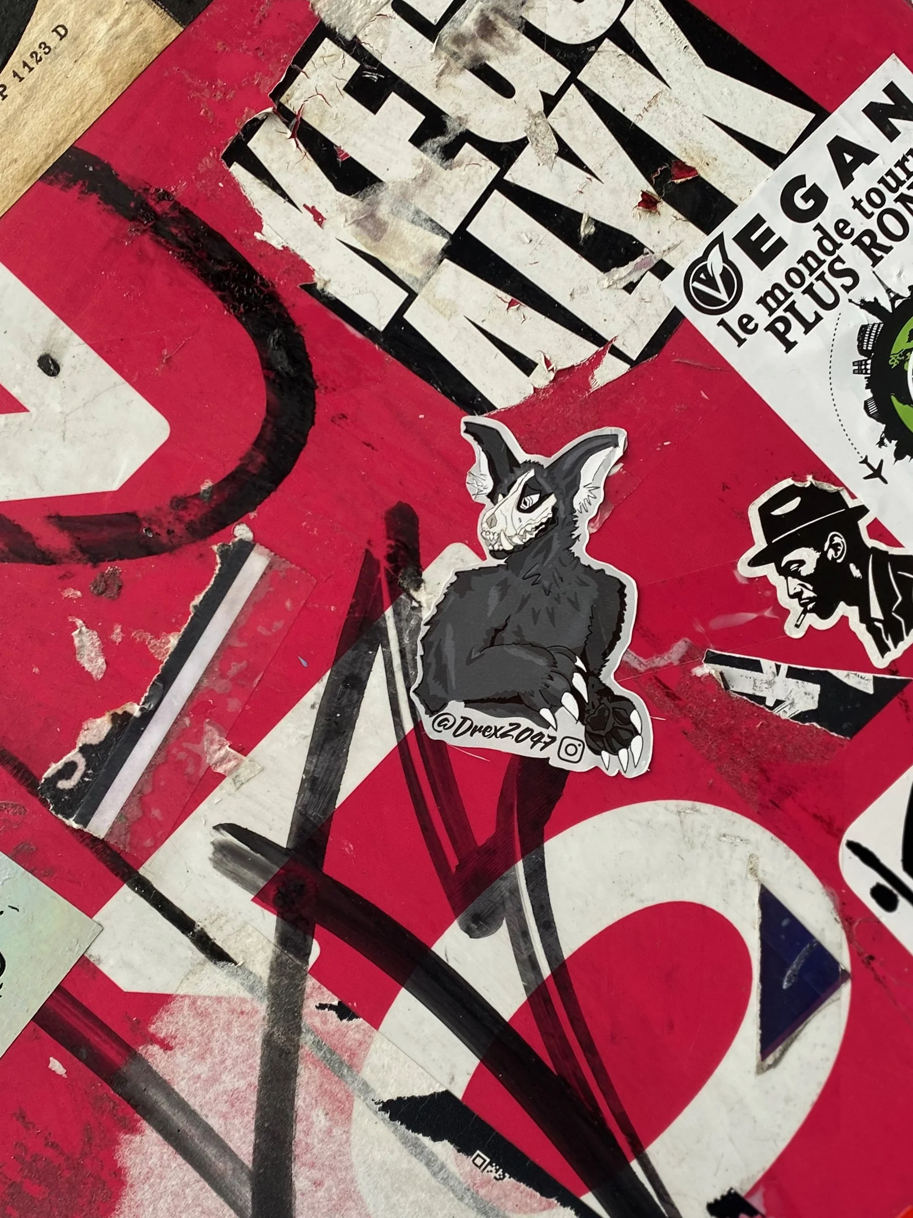 A sticker of a black and white cartoon creature with rabbit-like ears and a mischievous grin, sitting with crossed arms, on a red and white surface with other stickers and graffiti.