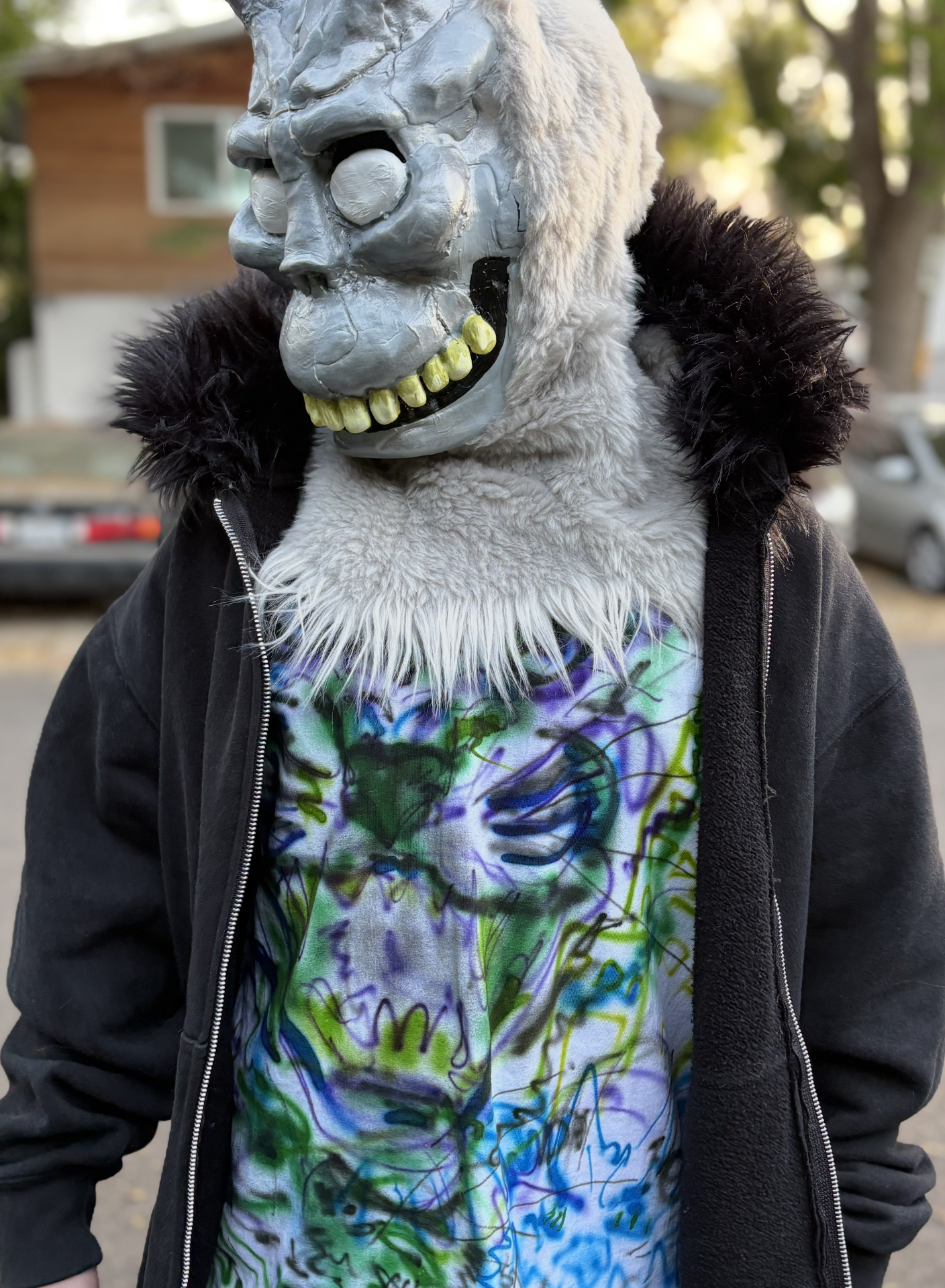 Person wearing a ghost skull mask with a black jacket and a colorful tie-dye shirt, standing outdoors with trees and a house in the background.