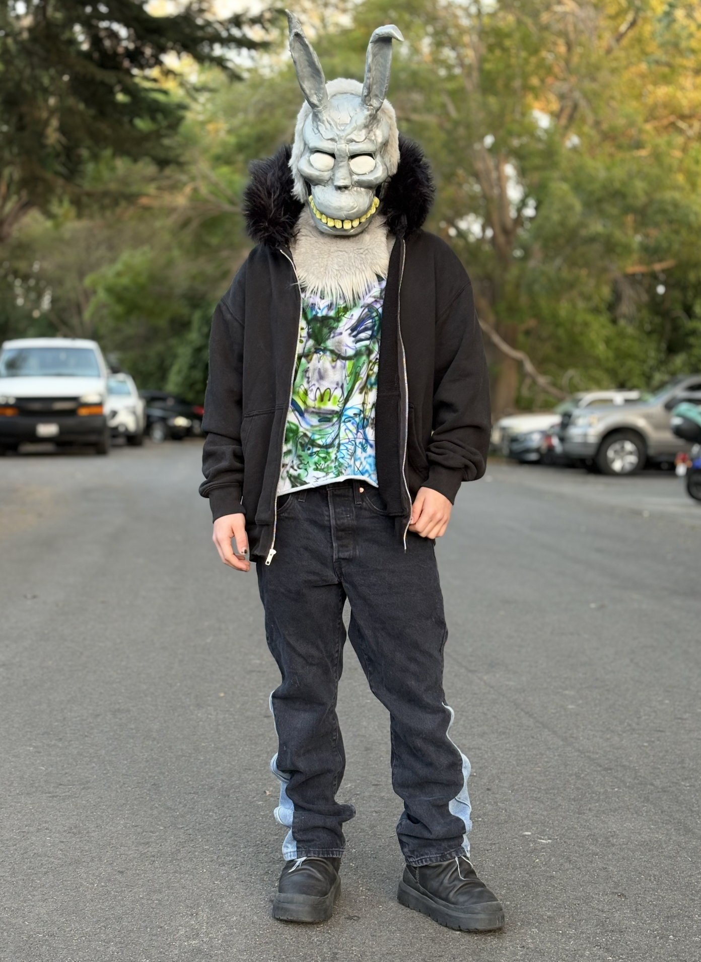 Person wearing a mask with a rabbit skull, large teeth, and long ears, dressed in a black jacket, graphic shirt, black jeans, and black boots, standing outdoors in a parking lot with trees in the background.