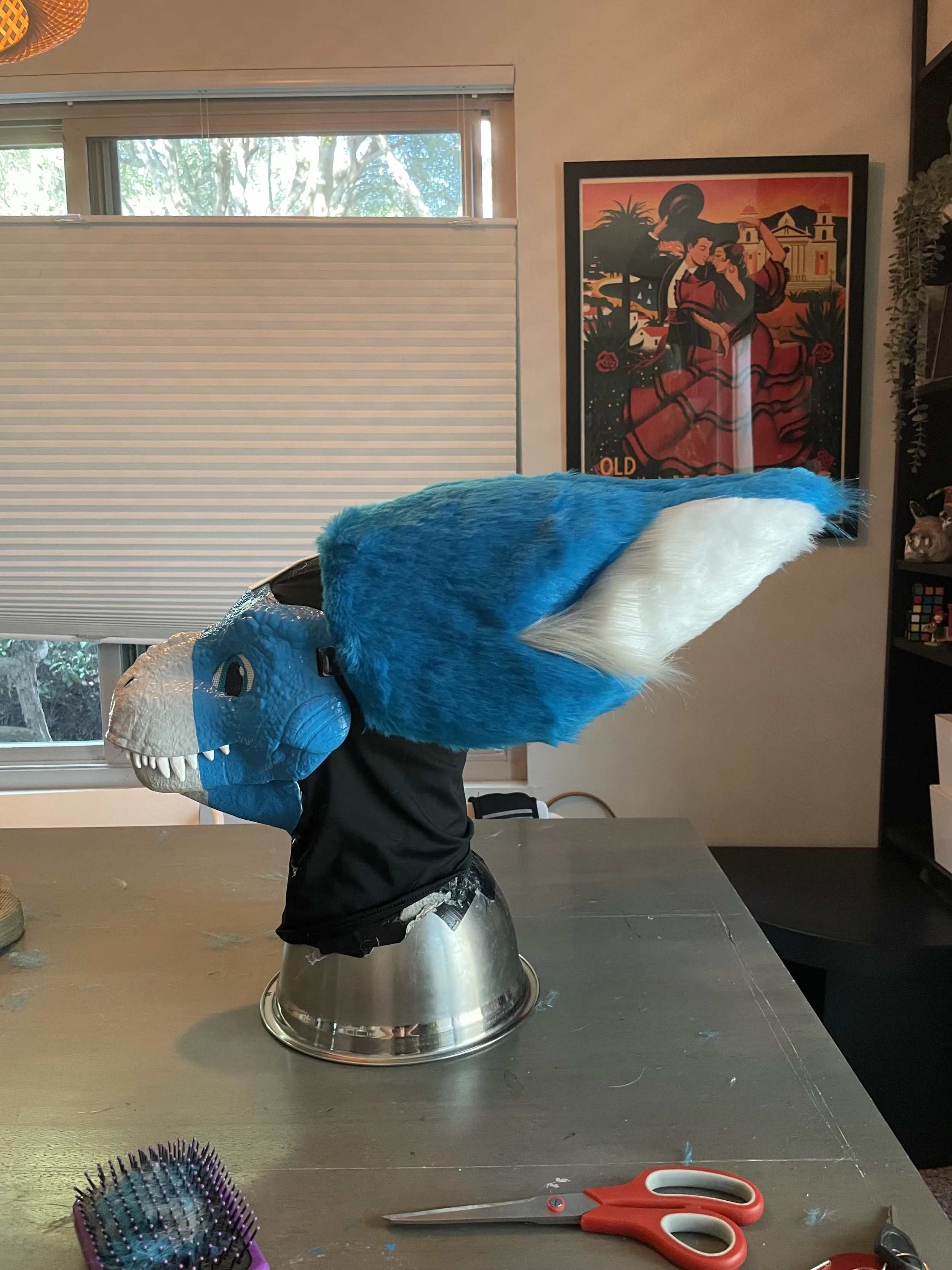 A mascot costume of a blue and white dragon head on a table, with scissors and a hairbrush nearby, and a window with a view of trees in the background.