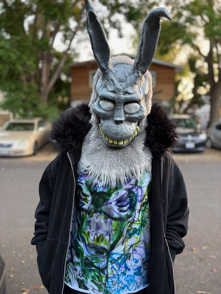 A person wearing a detailed gray rabbit or hare mask with large ears, a wide grin with yellow teeth, a furry neckpiece, a black jacket with a fur-lined hood, and a colorful, graffiti-style shirt, standing outdoors in a parking lot with trees and cars