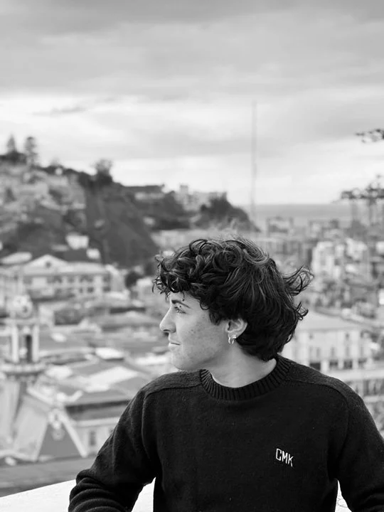 A young person with short curly hair and earrings, wearing a black sweater with 'CMK' on it, looking to the side against a cityscape background with buildings and hills in the distance.