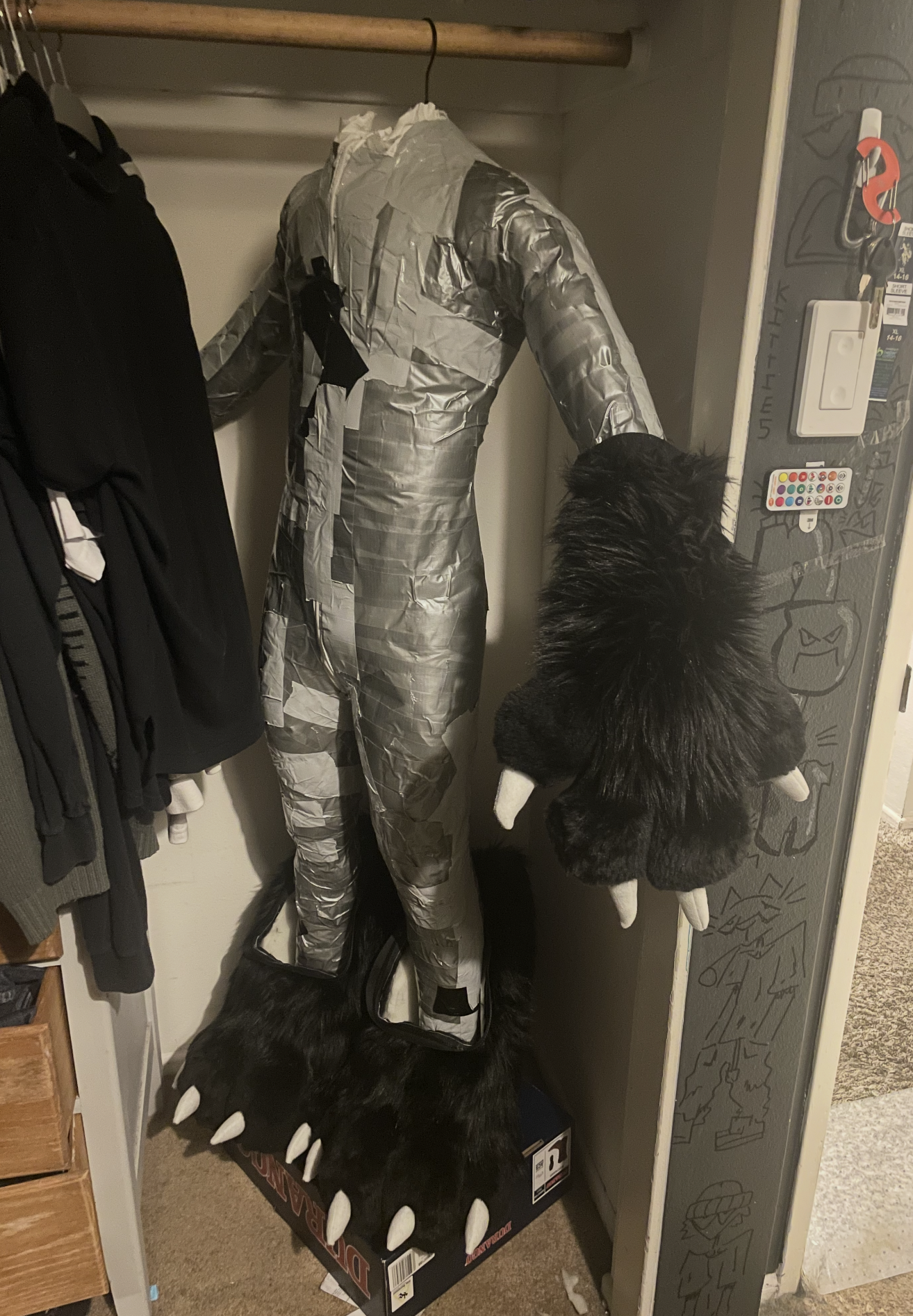 A mannequin dressed in a silver reflective outfit with a black harness, with black and white furry paws, and black furry parts at the bottom. The mannequin is in a closet next to hanging clothes, with a furry black and white headpiece at the bottom. 
