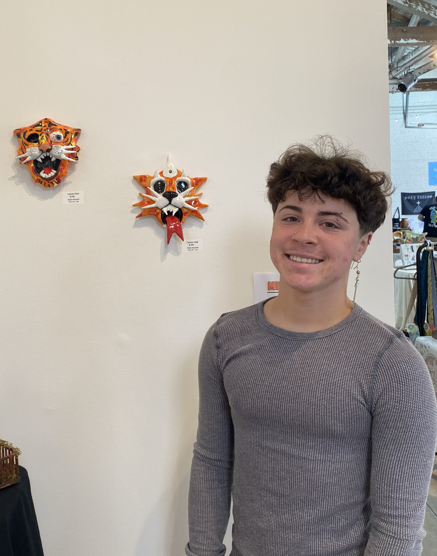 A young person with curly brown hair, smiling, wearing a gray long-sleeve shirt, standing in front of a white wall with two colorful ceramic masks, one tiger and one cat, displayed on the wall.