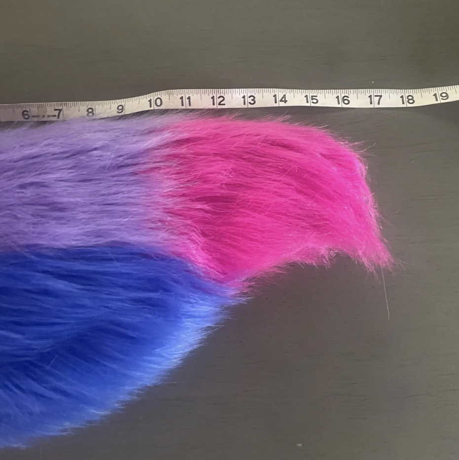 A colorful furry object with pink, purple, and blue fur on a dark surface, with a measuring tape above it.