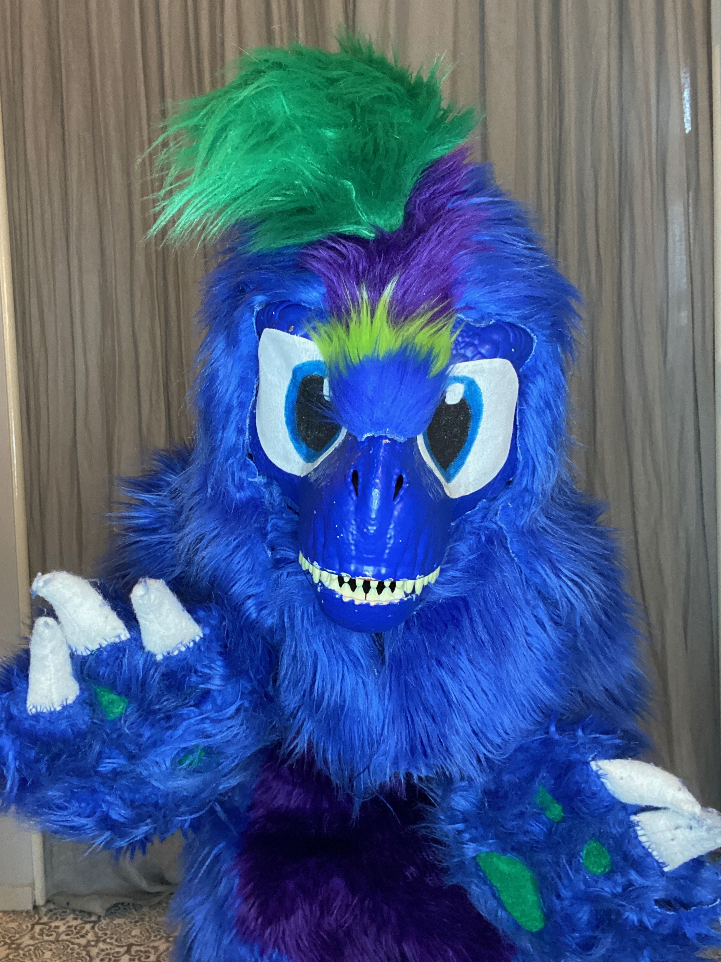 Person dressed in a colorful dinosaur costume with blue, purple, green, and white fur, large cartoonish eyes, and a toothy grin, waving at the camera.