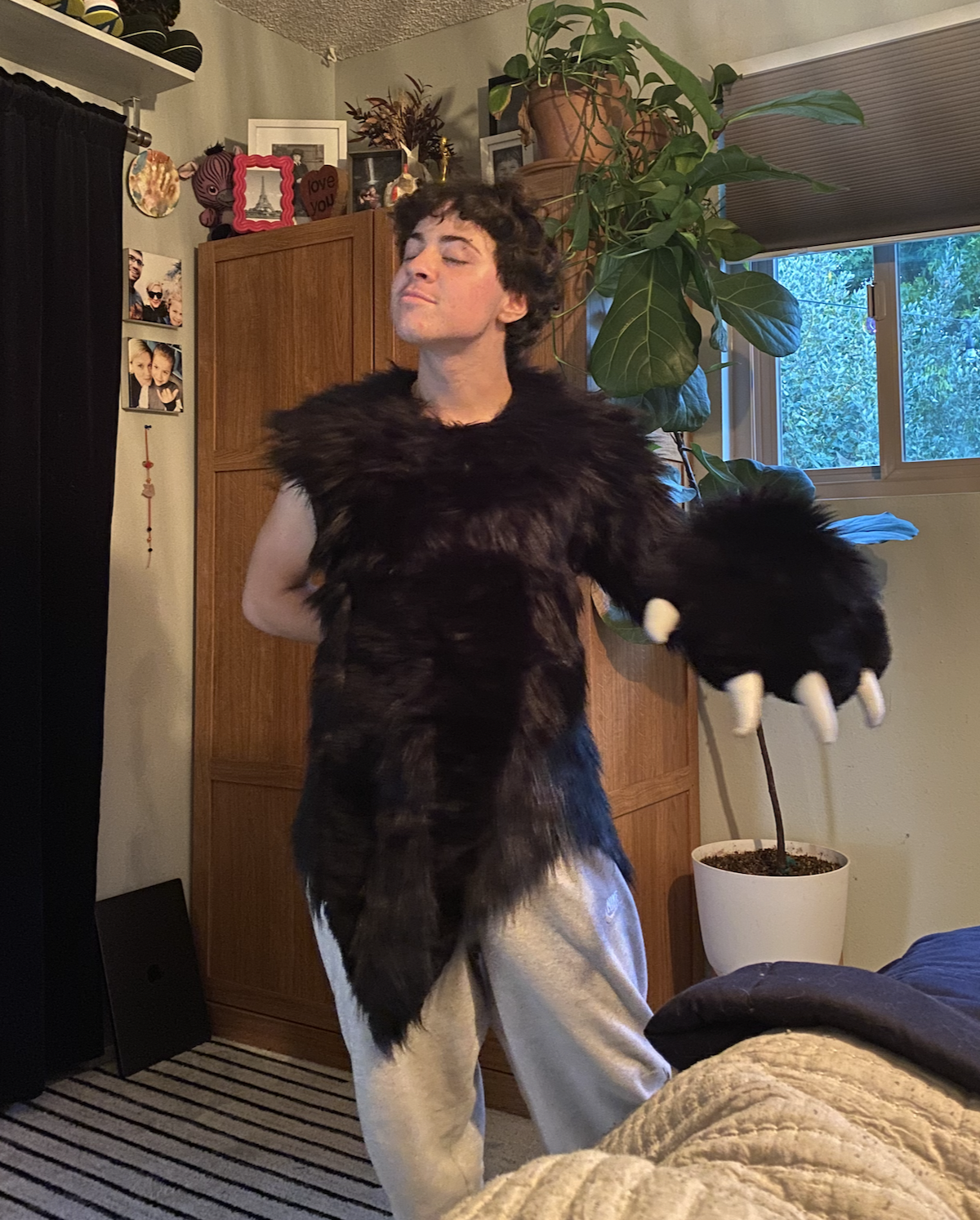 A person standing in a room wearing a black furry animal costume with large paws and claws, and a tail, with a large plant and pictures on the wall in the background.
