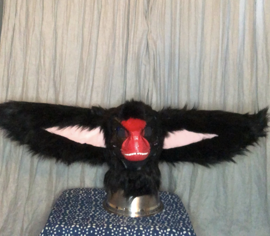 Furry animal mask with large ears and red nose mounted on a stand, placed on a blue polka dot cushion against a grey curtain background.