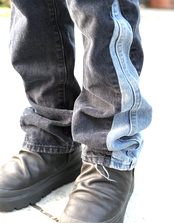 Close-up of a person's lower legs showing black boots, dark blue jeans with light blue stripes on the side, and a person standing outdoors on a concrete surface.