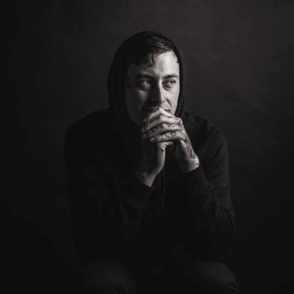 A black and white photo of a man wearing a hooded sweatshirt, sitting with his hands clasped near his face, looking thoughtfully to the side against a dark background.